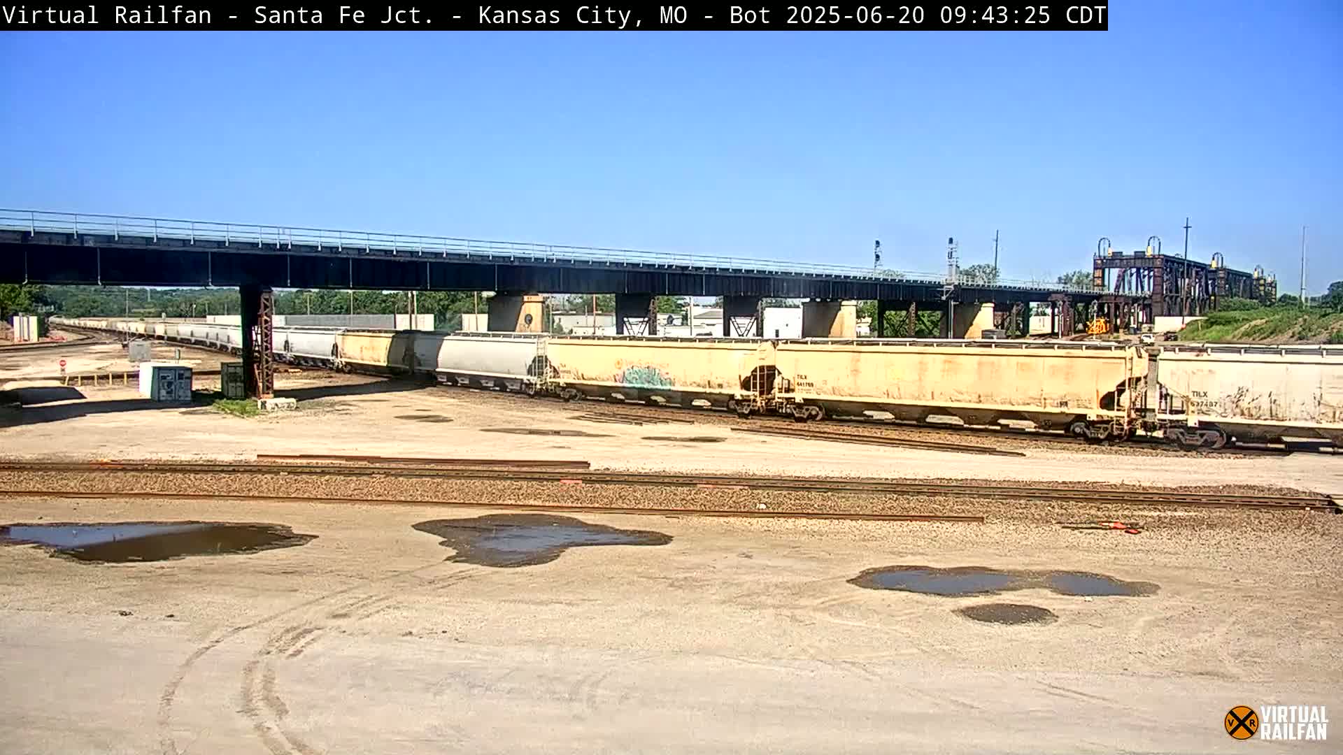 A sunny day reveals a rail yard with several light-beige covered hopper cars under a bridge, and several puddles on the ground.