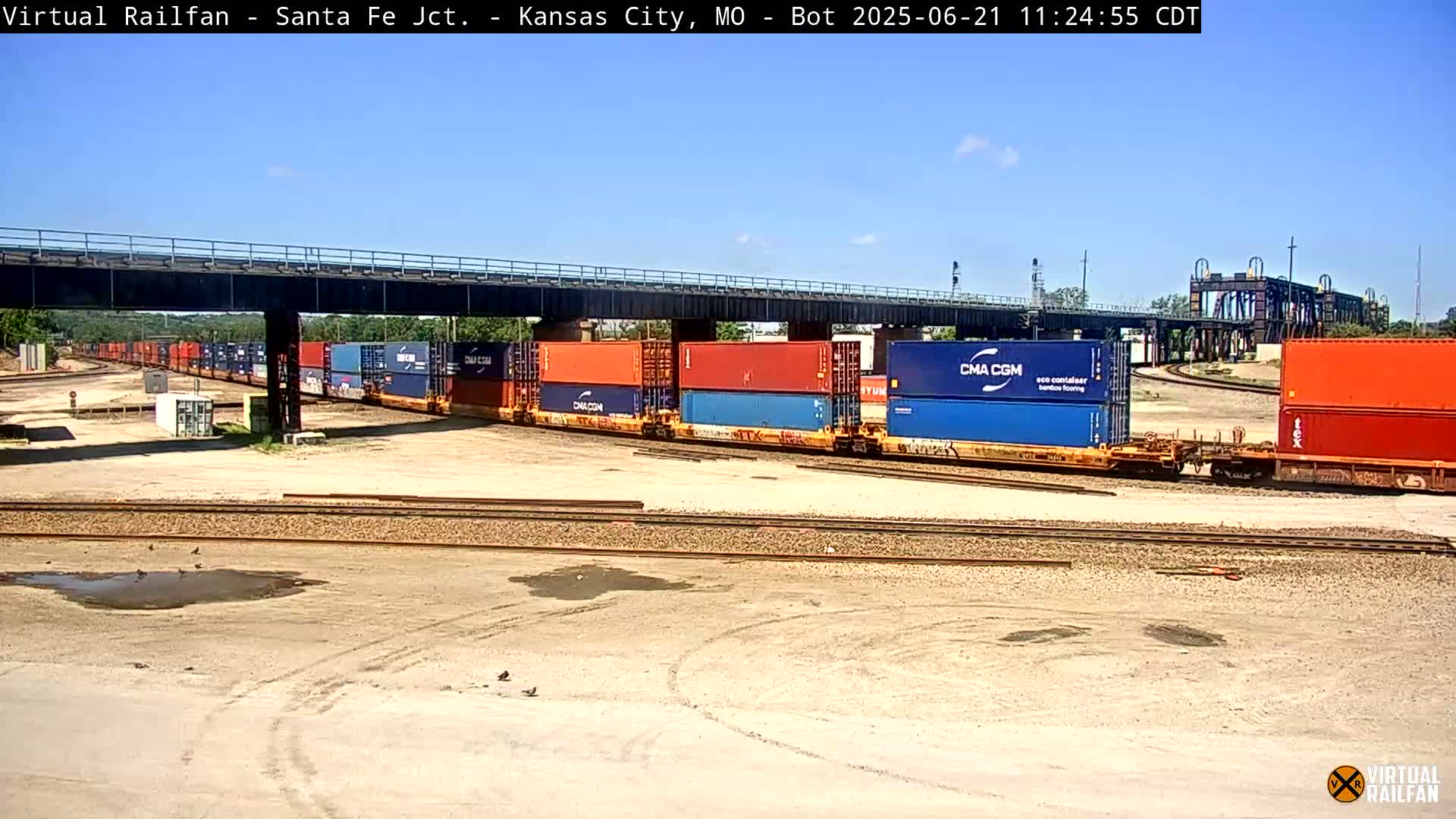 A long freight train carrying numerous shipping containers sits under a bridge on a sunny day in a rail yard.