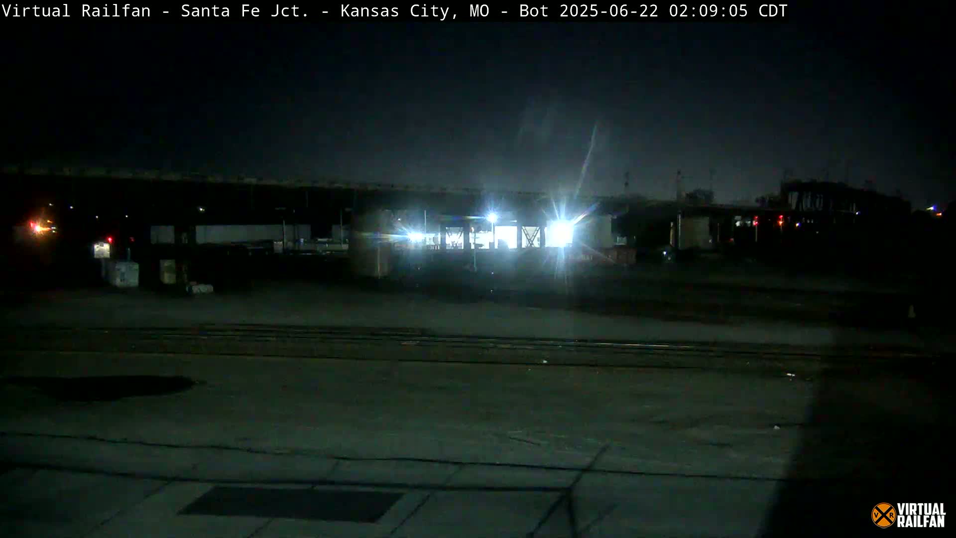 A nighttime, dimly lit train yard is shown with a train partially visible under a bridge.