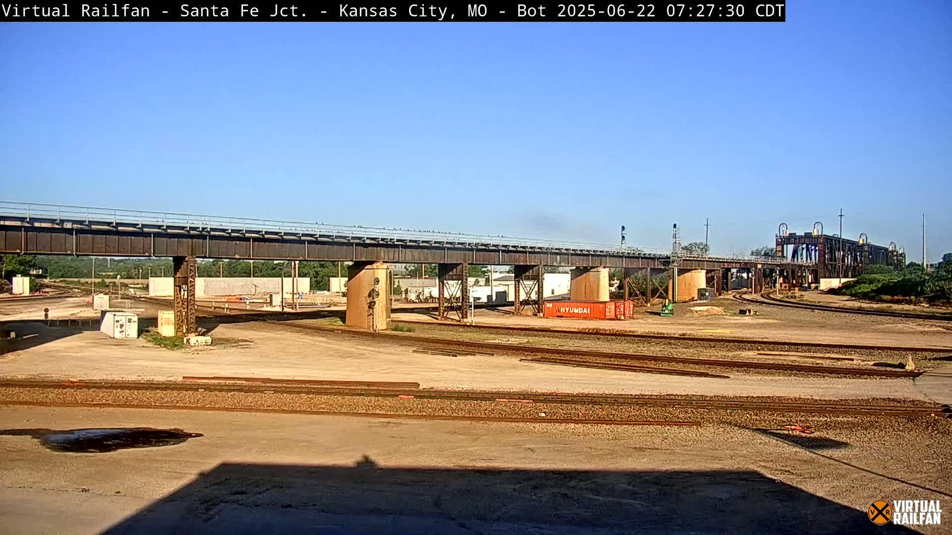 A railway yard with a large bridge overhead under a clear, sunny sky.
