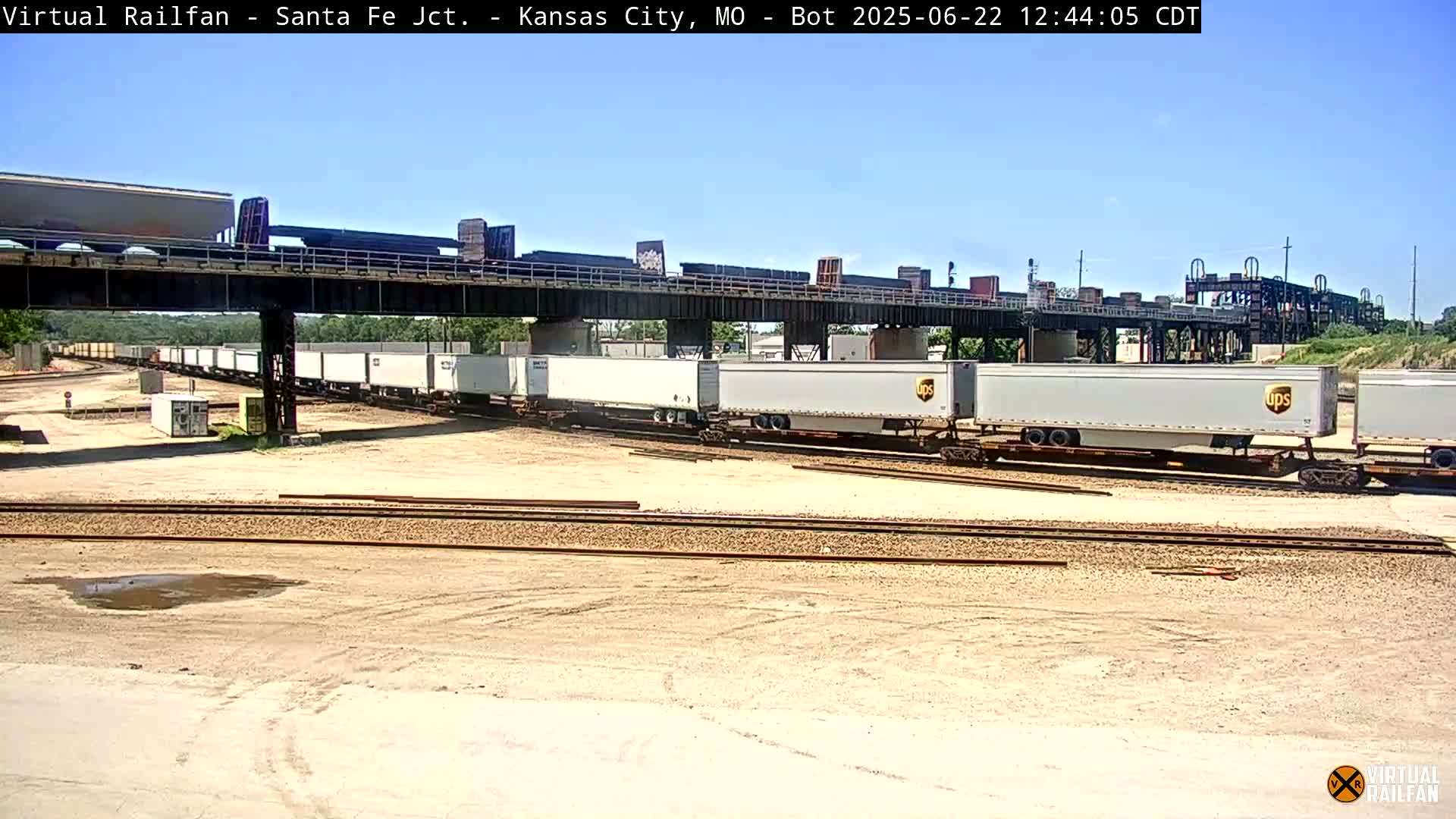 Under a clear blue sky, several long lines of UPS rail cars sit on train tracks below a bridge, surrounded by a flat, sandy area.