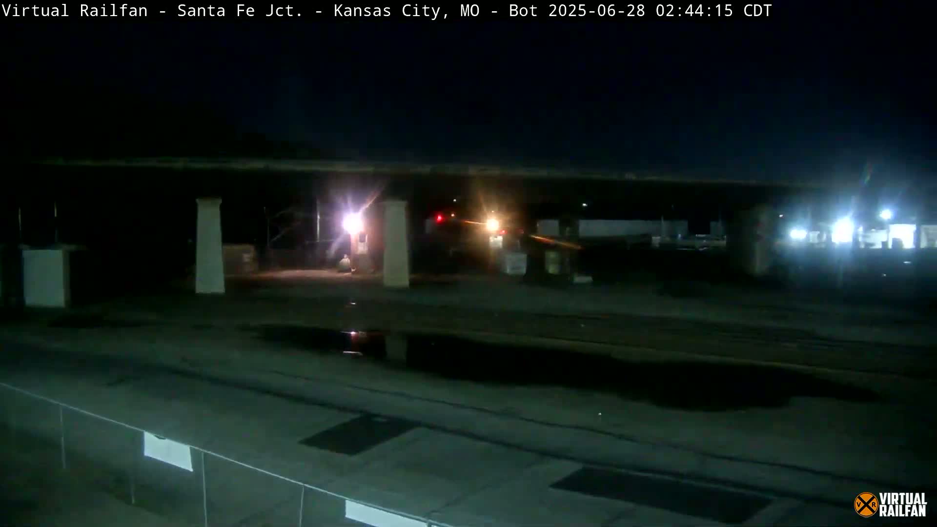 Under a dark night sky, several vehicles are visible near a structure with pillars, and a puddle reflects light on the wet ground.