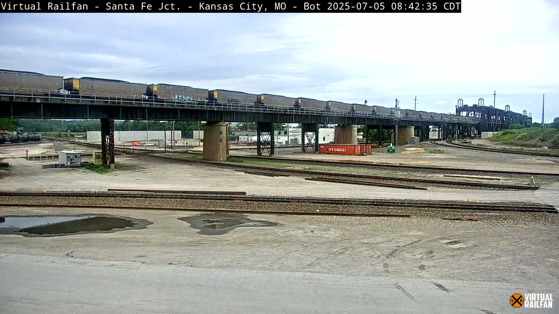 A long train of coal cars crosses a railway overpass on an overcast day in a rail yard.