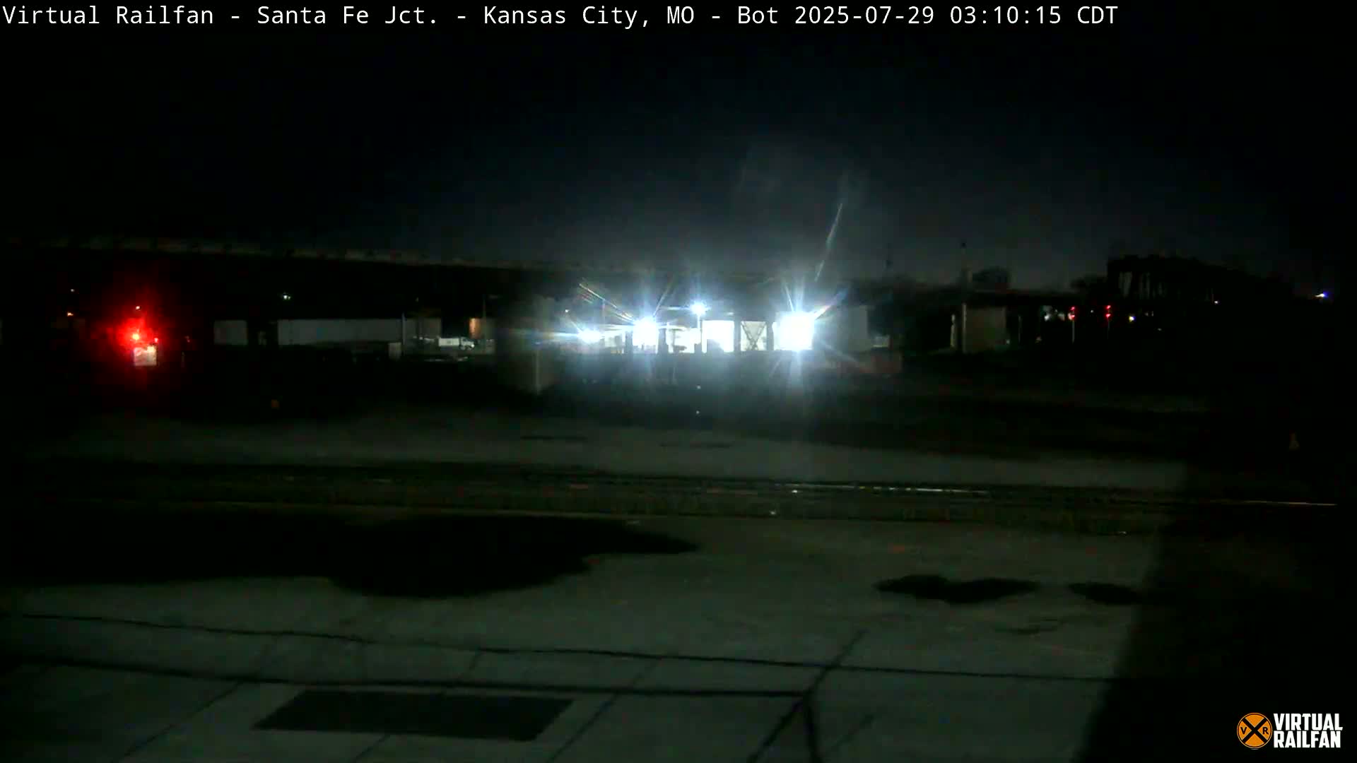 A nighttime view of a train illuminated by bright lights, passing under a structure in a dimly lit area.