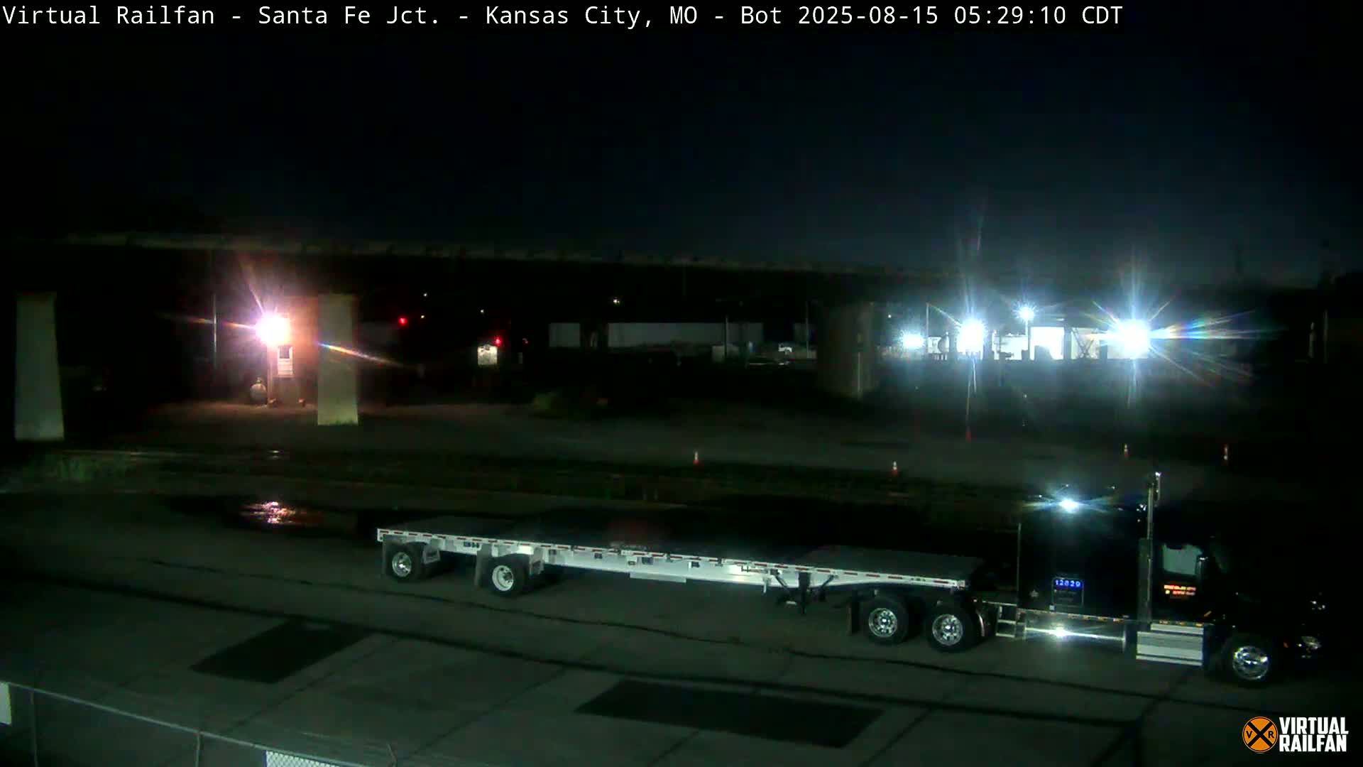A nighttime view of a train illuminated by bright lights, passing under a structure in a dimly lit area.