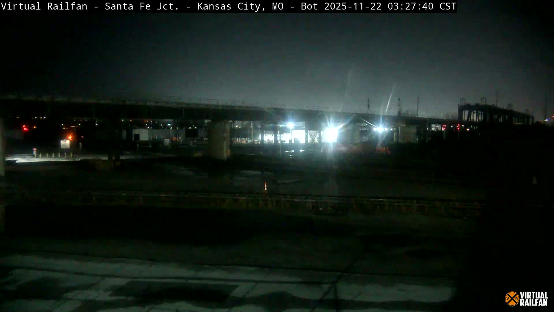 The image shows a damp, overcast night in an industrial or construction area beneath an overpass, where bright artificial lights reflect on wet ground.