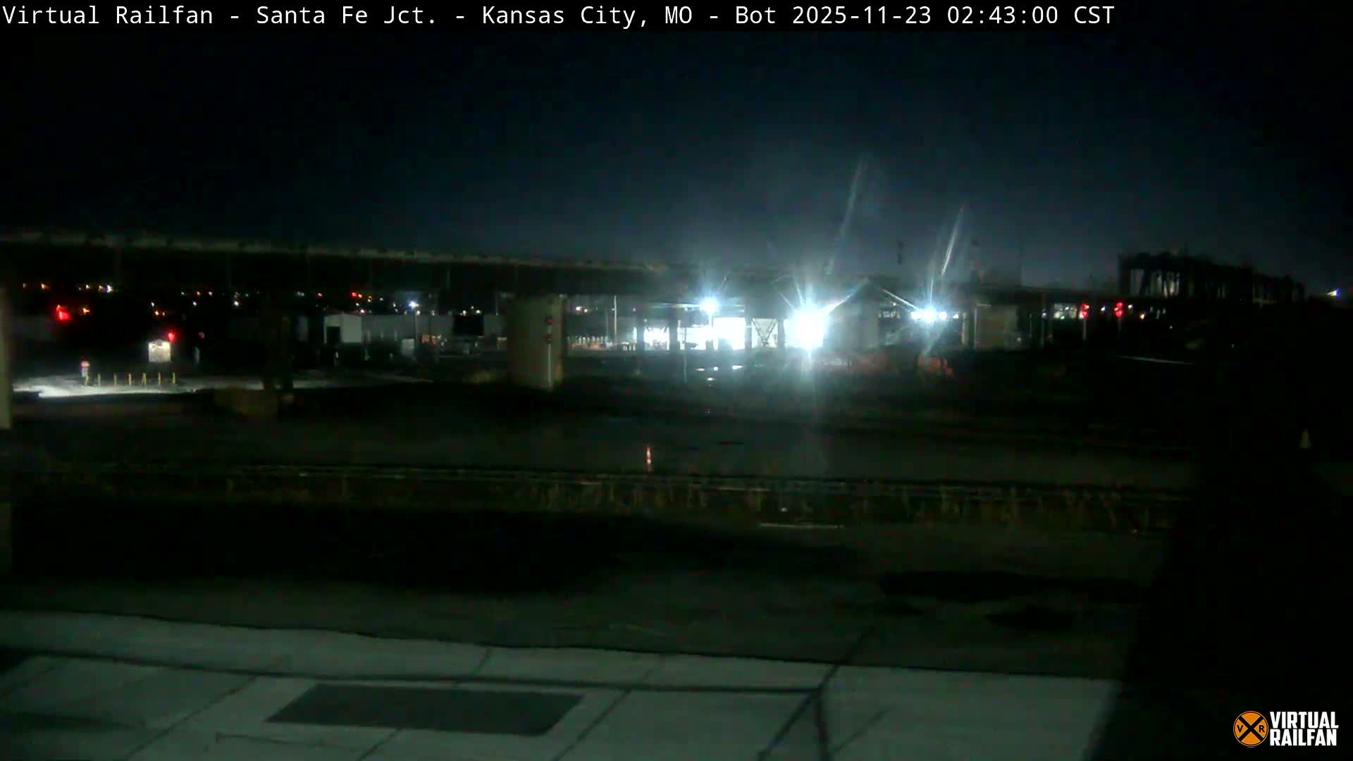 A clear, dark night scene illuminates a busy railway junction or industrial area, featuring multiple tracks in the foreground, an overhead structure supported by pillars, and various buildings brightly lit by artificial lights in the midground.