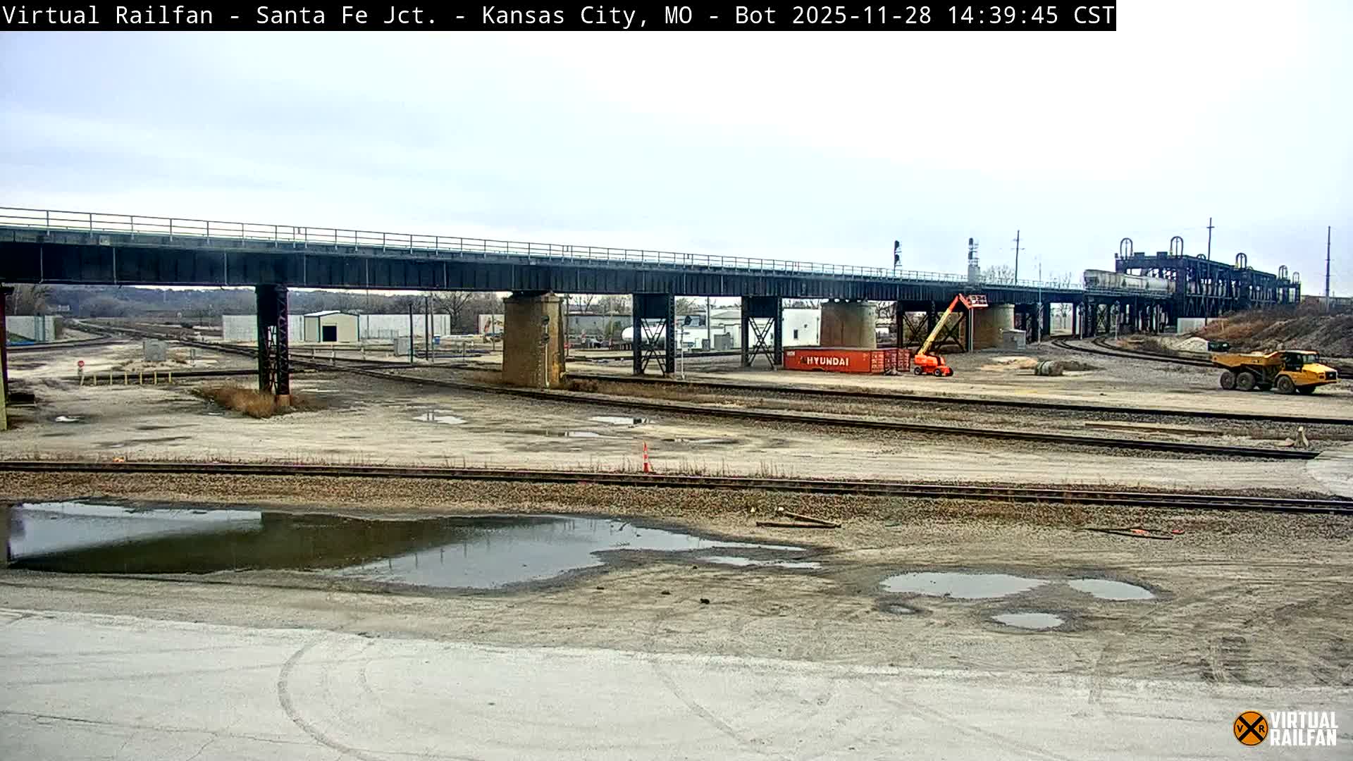 An expansive view of an industrial railyard under an overcast sky features a prominent metal railway bridge crossing multiple tracks, with numerous puddles on the dirt ground and a construction lift alongside a large yellow dump truck.
