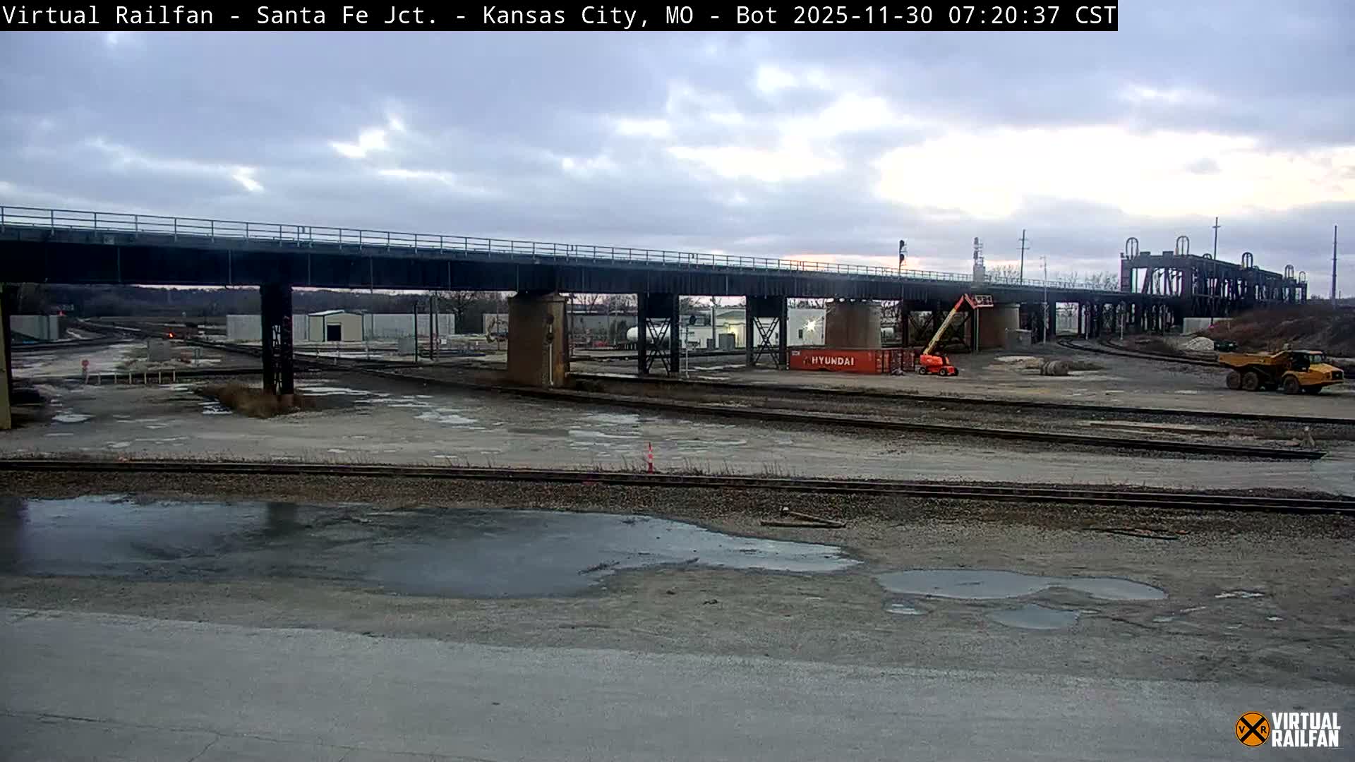 An expansive view of an industrial railyard under an overcast sky features a prominent metal railway bridge crossing multiple tracks, with numerous puddles on the dirt ground and a construction lift alongside a large yellow dump truck.