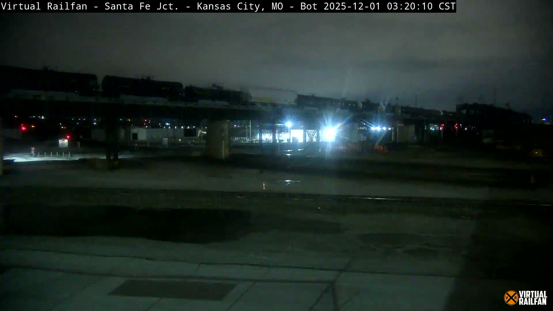 A long train is seen on an elevated track above ground-level railway lines and industrial buildings at night, with wet ground reflecting bright lights under an overcast sky.