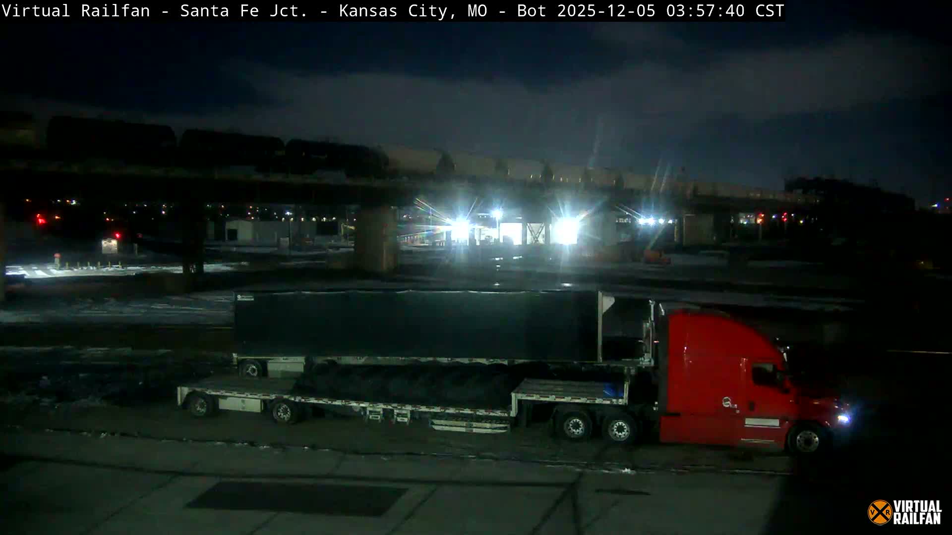 A red semi-truck with two flatbed trailers, one loaded with large coils and the other empty, is seen on a clear, dark, and likely cold night, beneath an elevated railway bridge carrying a train, adjacent to a brightly lit industrial or rail yard.
