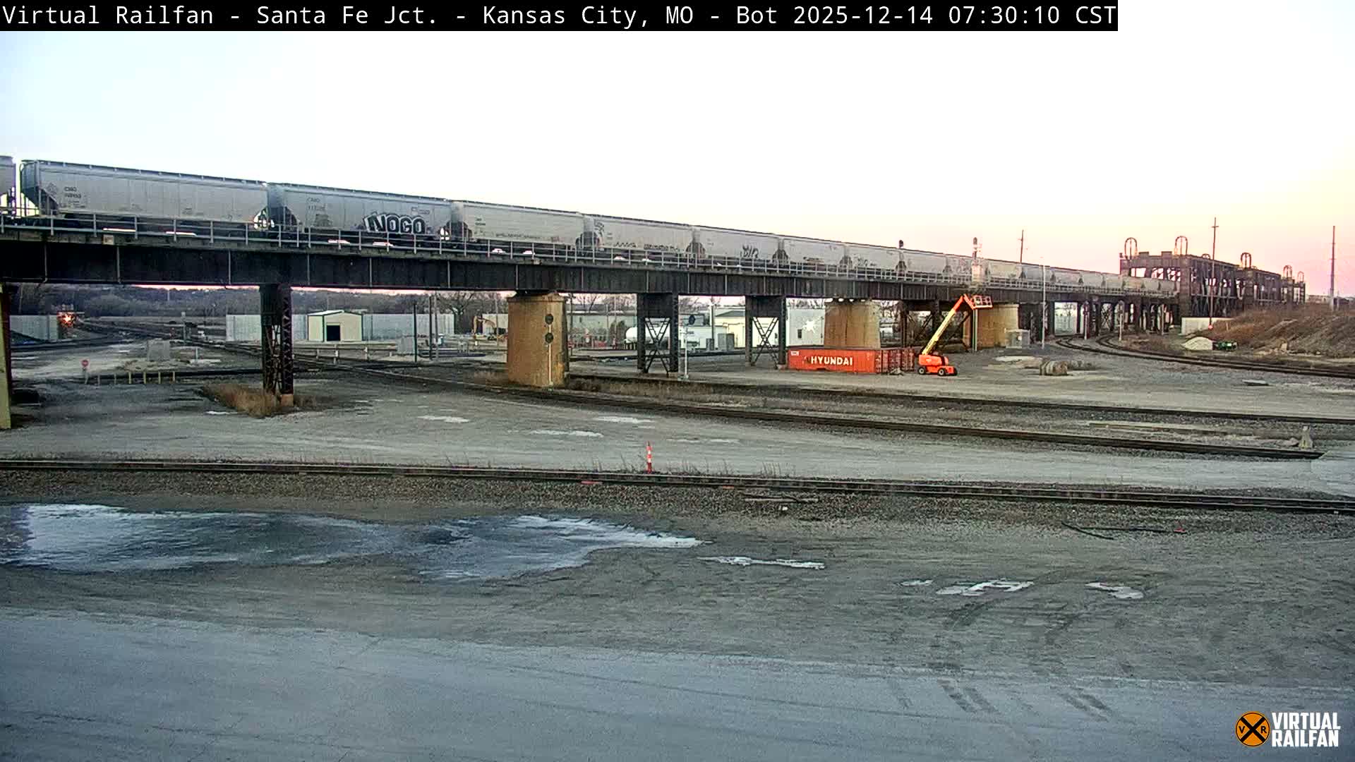 A red semi-truck with two flatbed trailers, one loaded with large coils and the other empty, is seen on a clear, dark, and likely cold night, beneath an elevated railway bridge carrying a train, adjacent to a brightly lit industrial or rail yard.