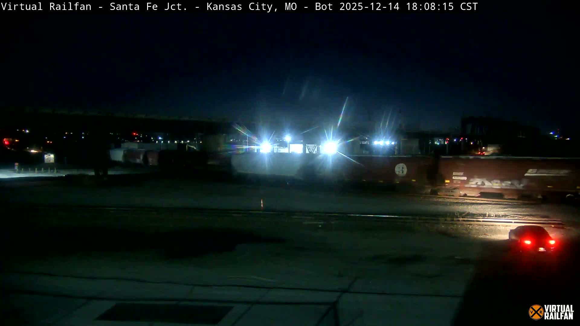 A red semi-truck with two flatbed trailers, one loaded with large coils and the other empty, is seen on a clear, dark, and likely cold night, beneath an elevated railway bridge carrying a train, adjacent to a brightly lit industrial or rail yard.