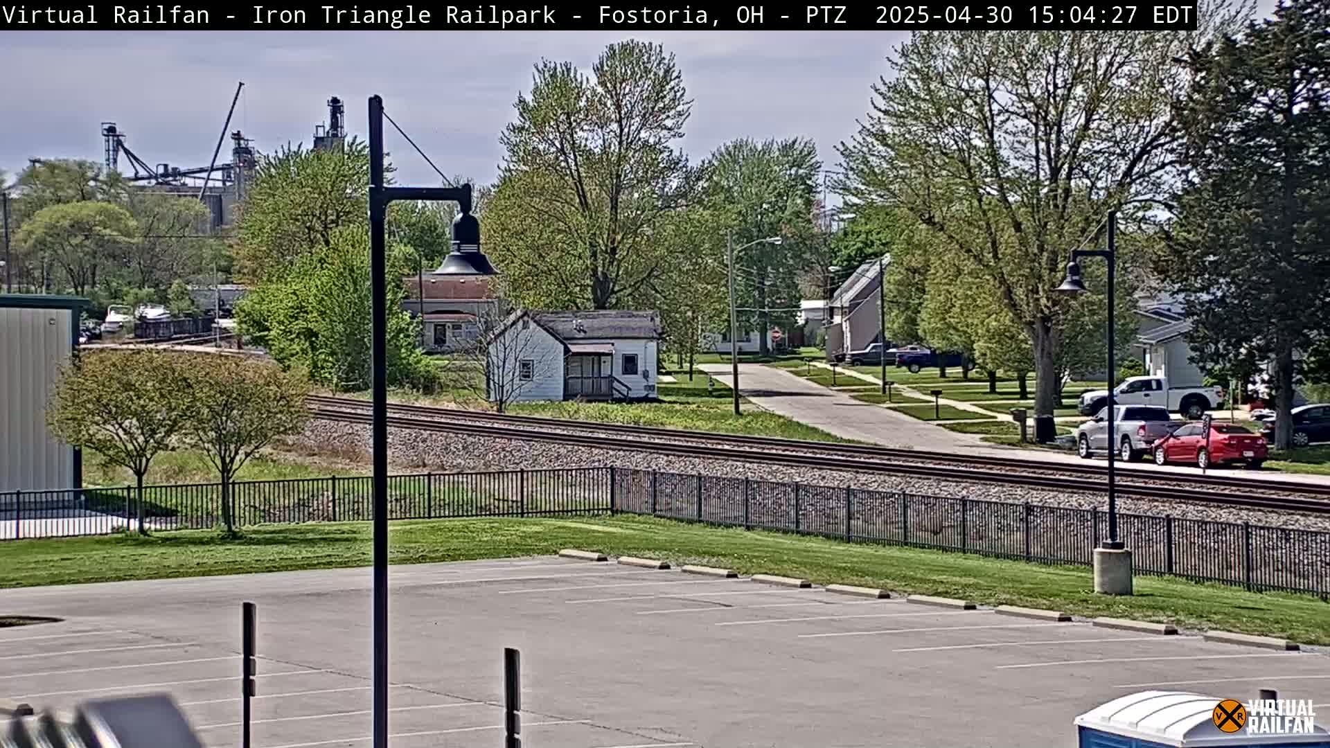 A view of a residential street, railroad tracks, and an industrial area in the background on a partly sunny day.