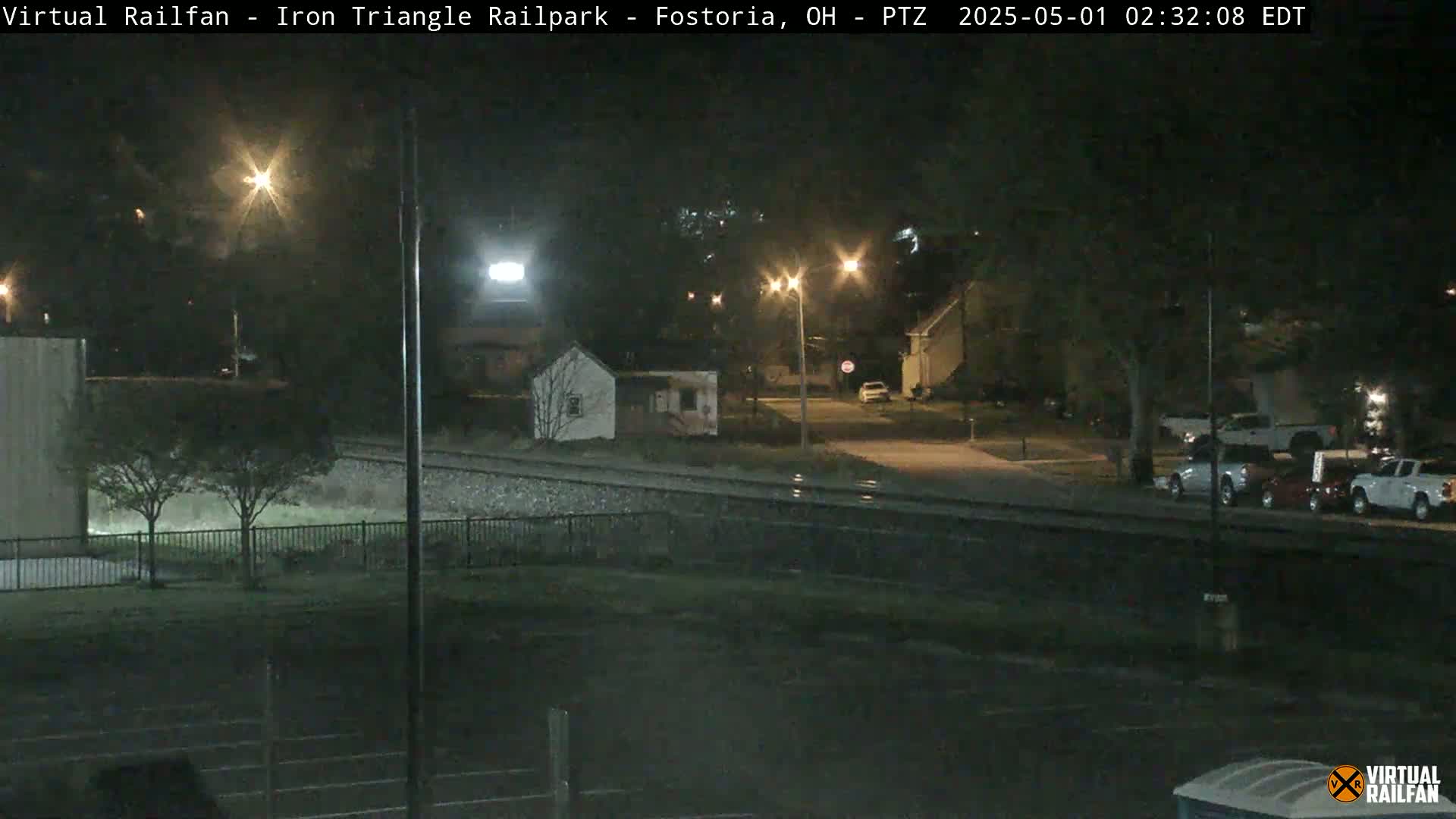 A nighttime view of a residential area near railroad tracks, with several parked cars and streetlights illuminating the scene.