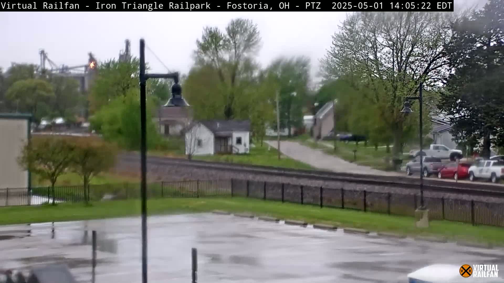 A rain-soaked parking lot and a view of a small town with houses and a rail line, beyond which are industrial structures and trees.