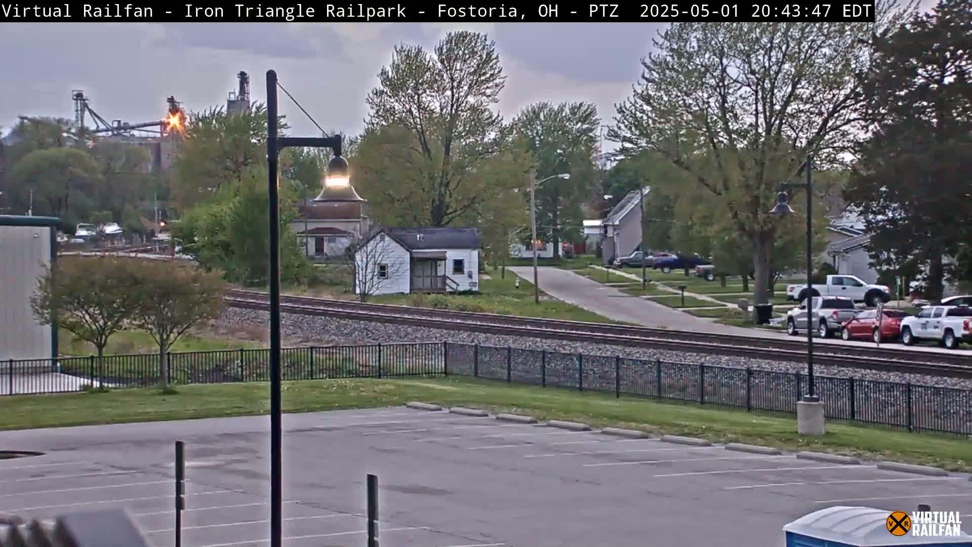 A mostly empty parking lot overlooks a railroad track bordering a residential street under a twilight sky.