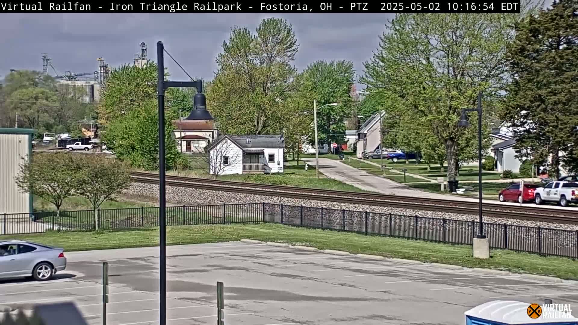 A mostly sunny day shows a residential area with a parking lot, railroad tracks, and a few cars, all surrounded by leafy green trees.