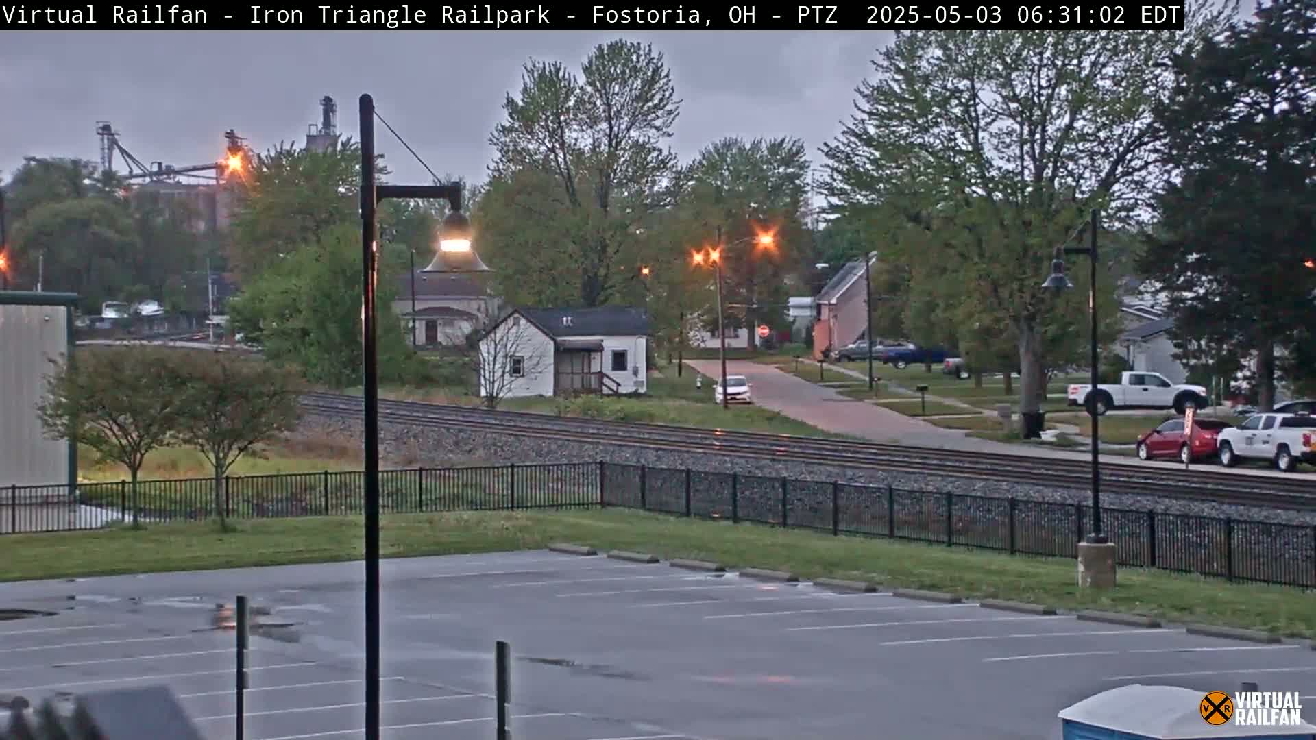A cloudy early morning scene shows a parking lot, railroad tracks, a small residential area with cars parked along the road, and industrial buildings in the background.