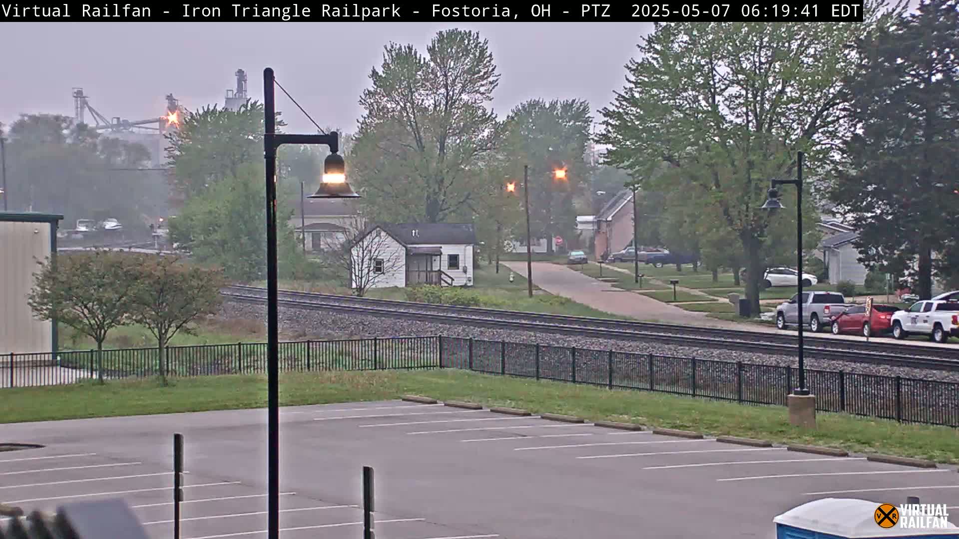 A misty view of railroad tracks alongside a residential street, with cars parked on the street and an empty parking lot in the foreground.
