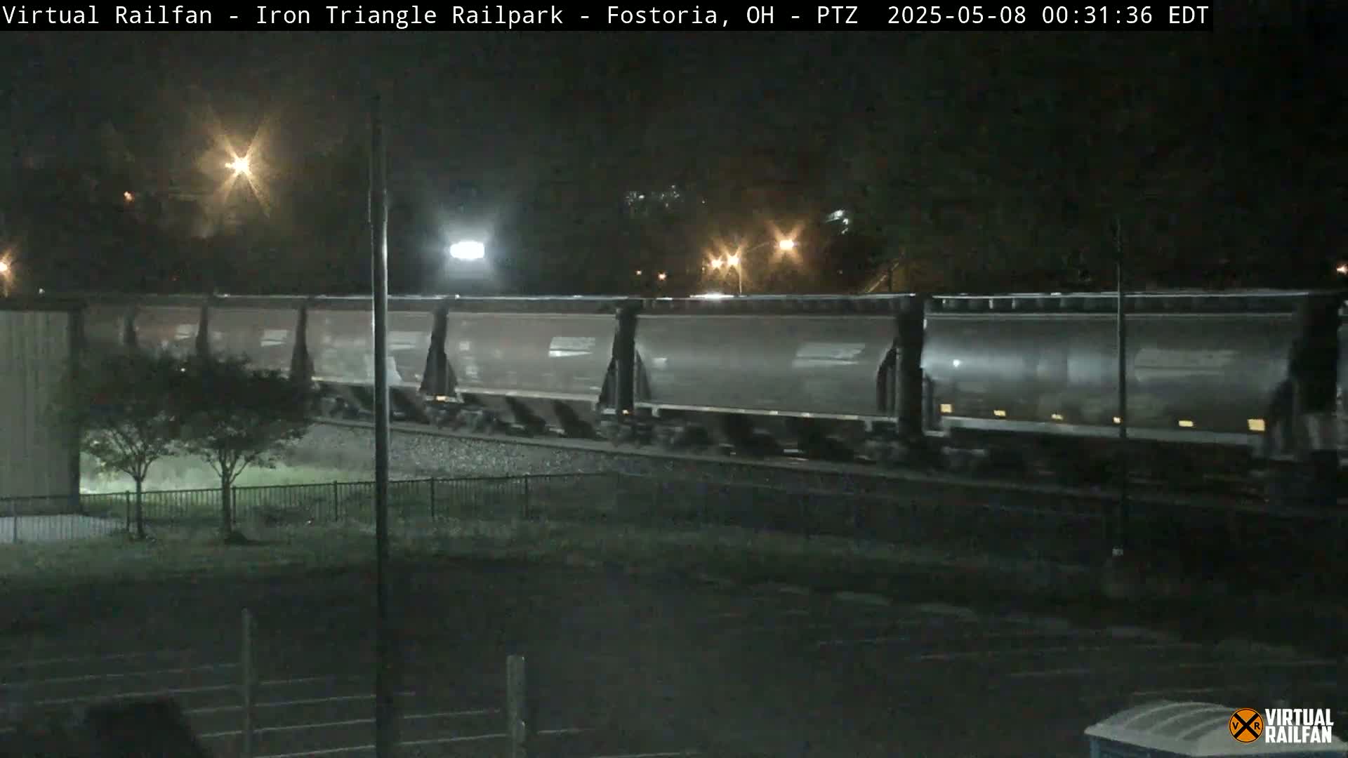 A nighttime view of a freight train moving along tracks, under artificial lighting.