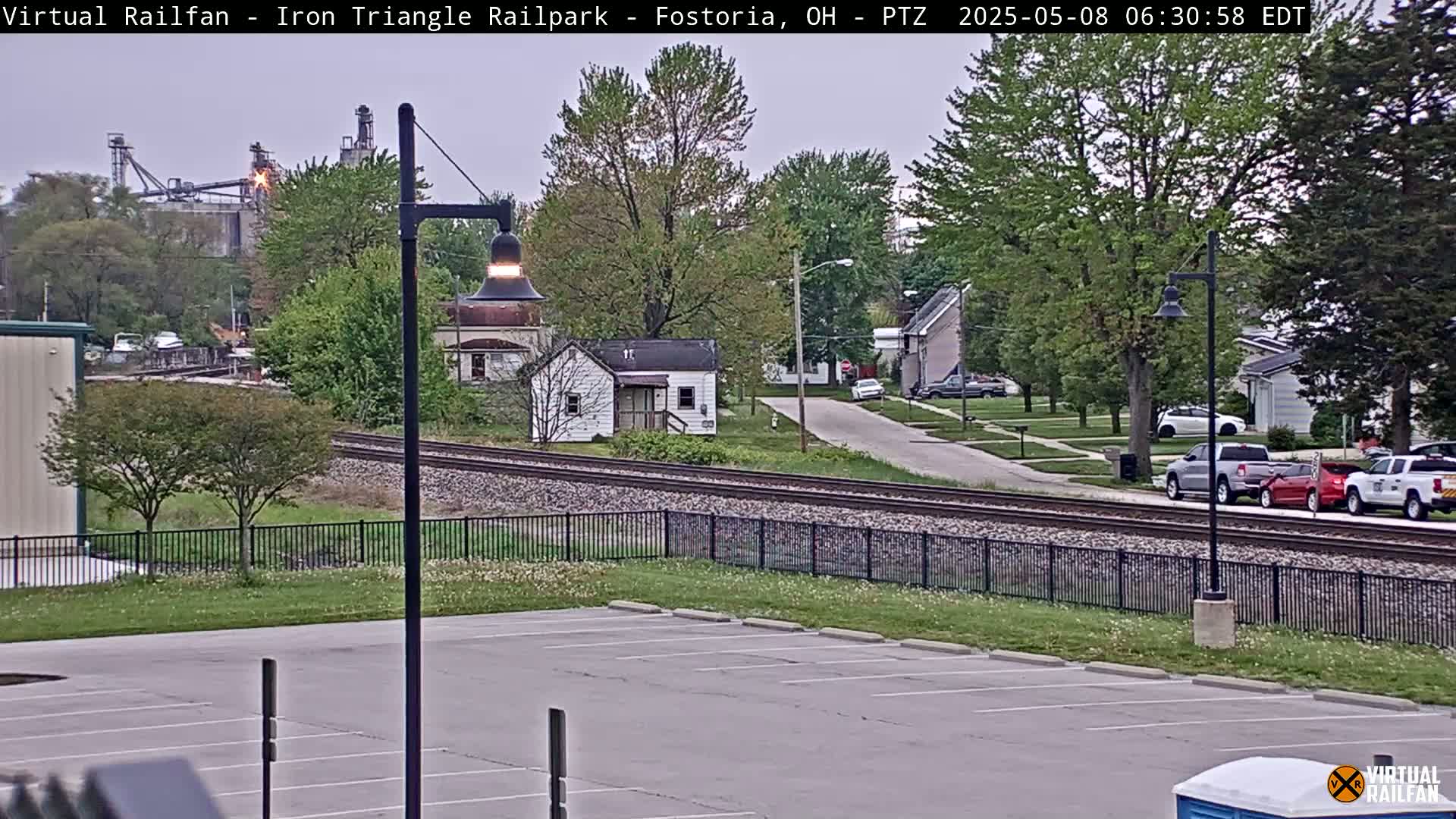 A view of railroad tracks beside a residential street on an overcast day, with a factory visible in the background.