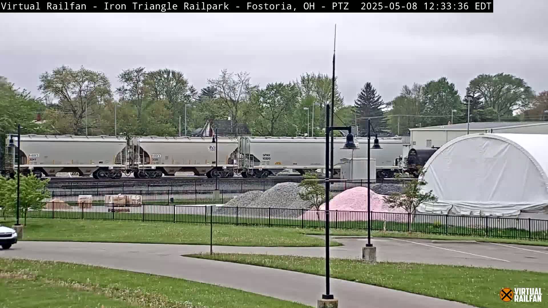 A freight train of light gray hopper cars sits on railroad tracks behind a chain-link fence, next to piles of gray and pink gravel under an overcast sky.
