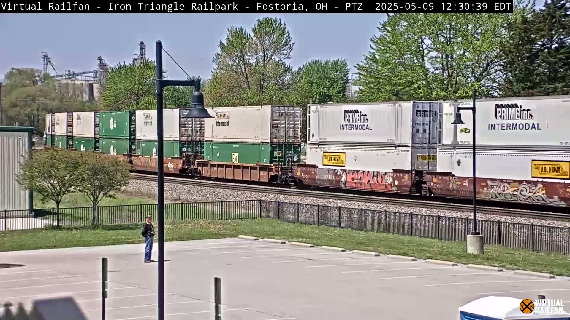 A long freight train carrying multiple shipping containers passes by a person standing in an empty parking lot on a sunny day.