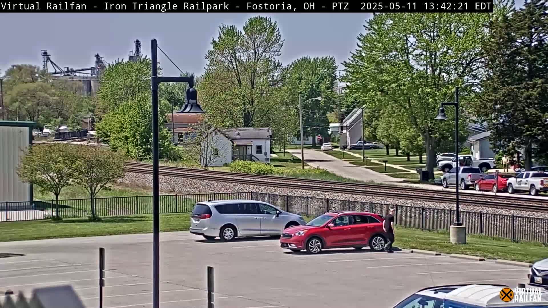 A sunny day shows a parking lot with a silver minivan and a red SUV parked, near railroad tracks with a person walking by, and a residential street beyond, with industrial buildings in the distance.