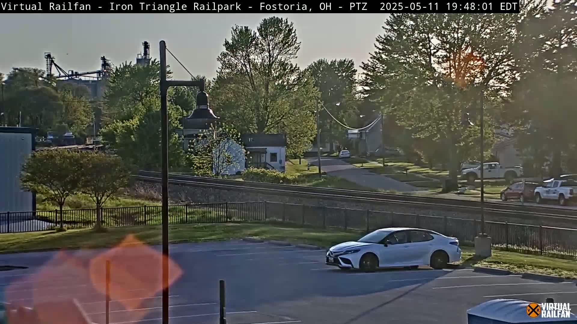A white sedan is parked in a lot next to railroad tracks, with a small building and houses visible in the background under a partly sunny evening sky.