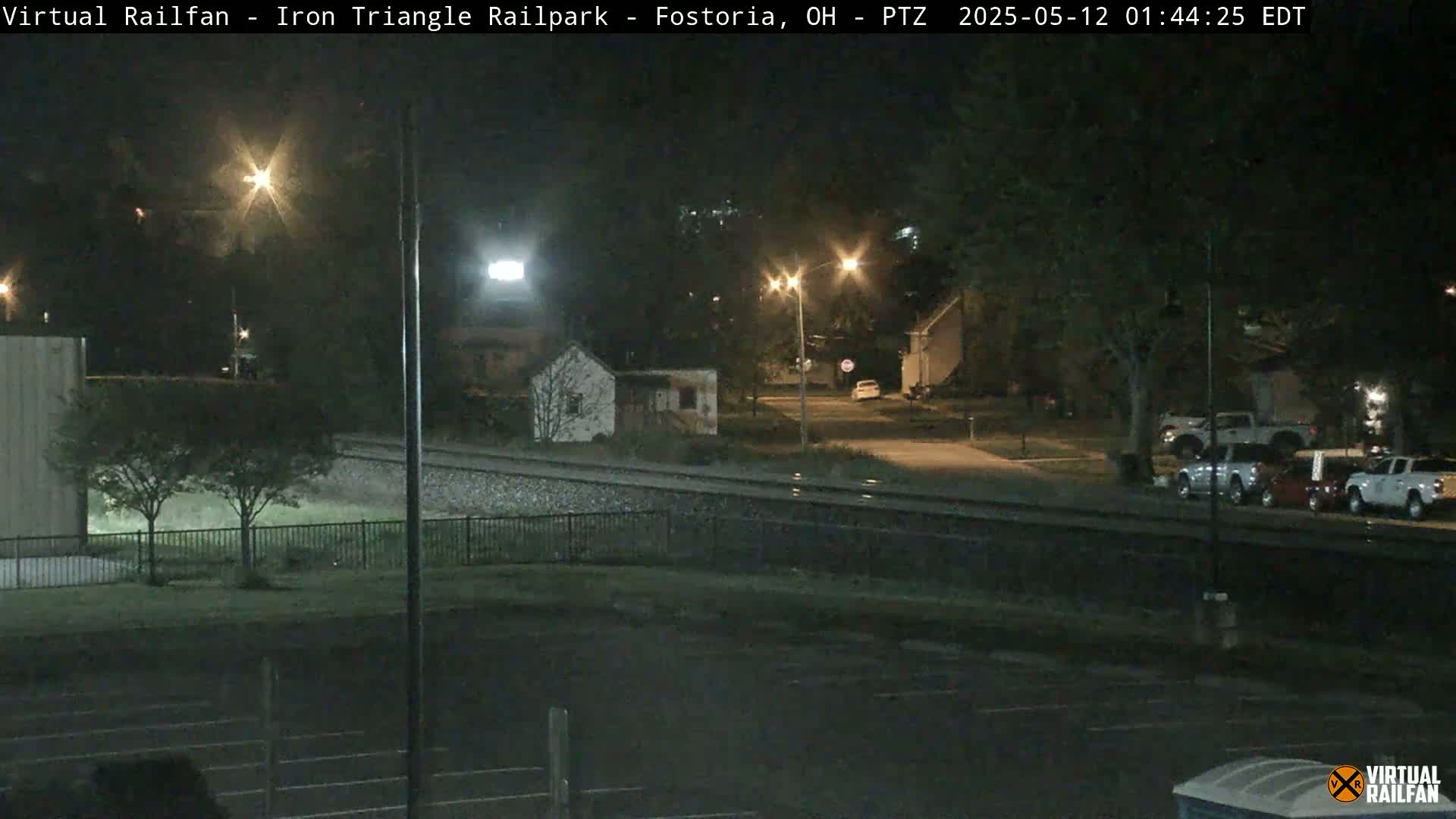 A nighttime view of a rail yard with several parked vehicles and a small building near a street, under clear skies.