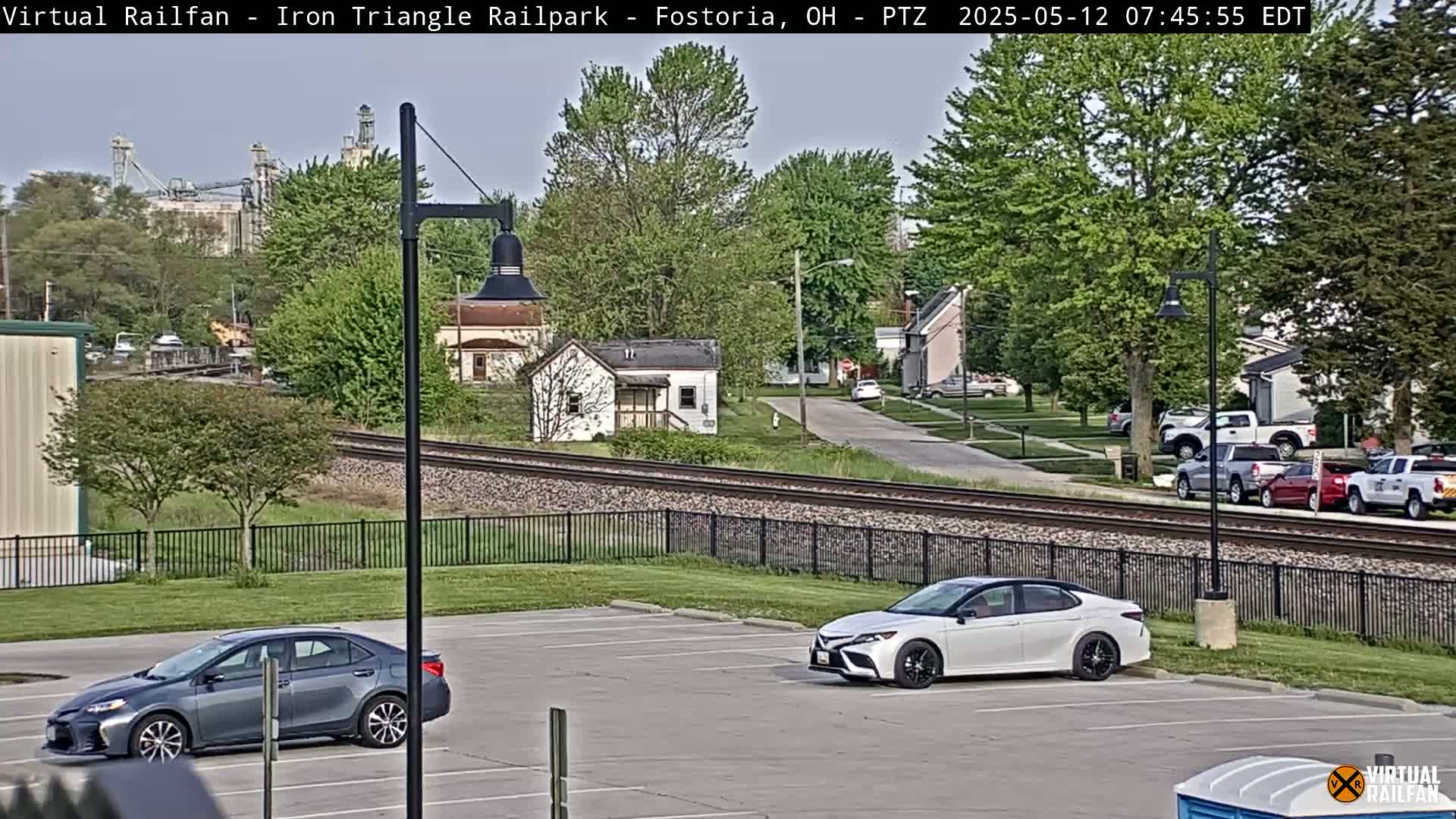 A parking lot with two cars, next to railroad tracks that run beside a residential street with parked cars and a grain silo in the distance, on a partly sunny day.