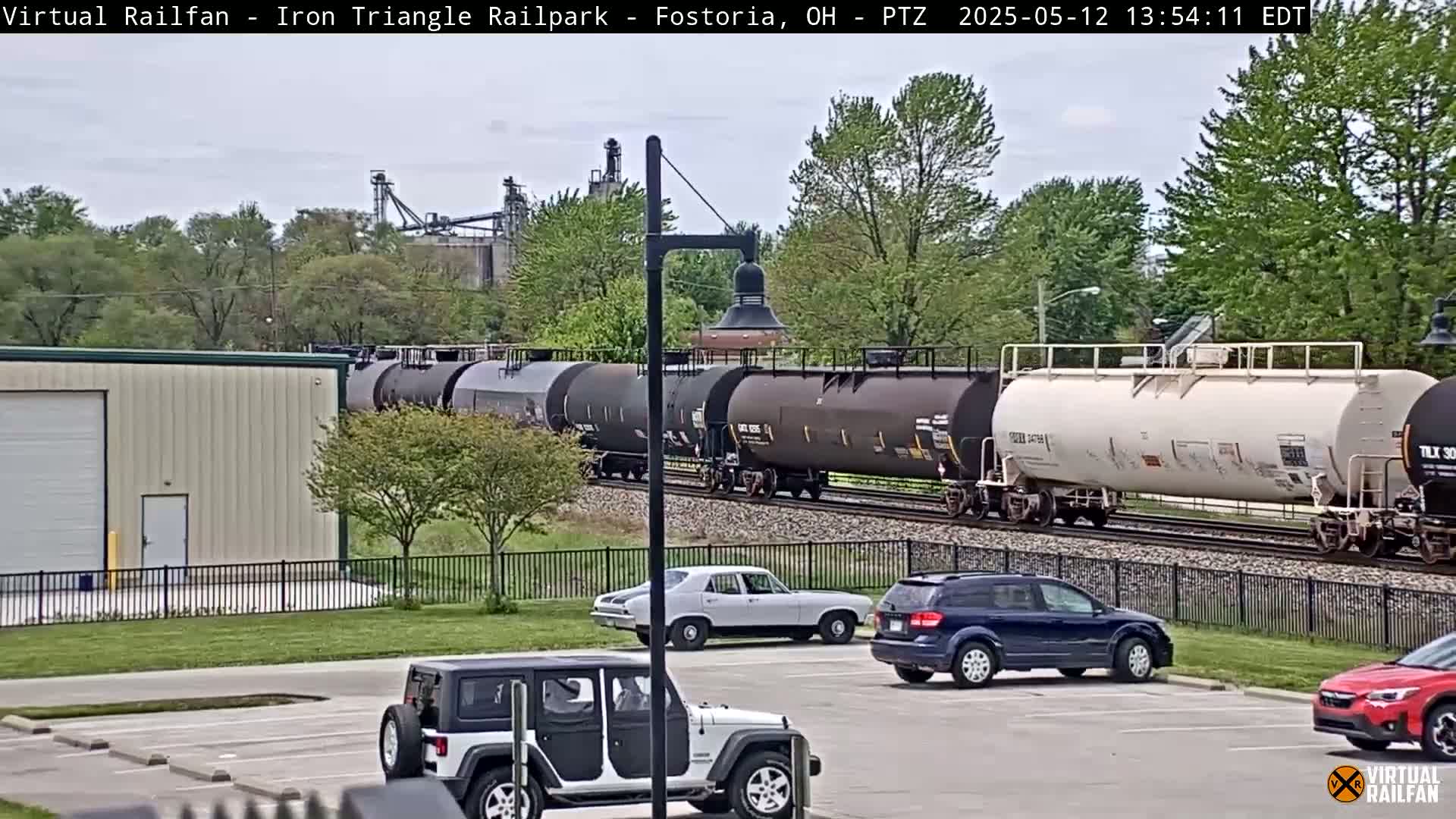 A long freight train passes by a parking lot with several cars and a jeep, under a partly cloudy sky.