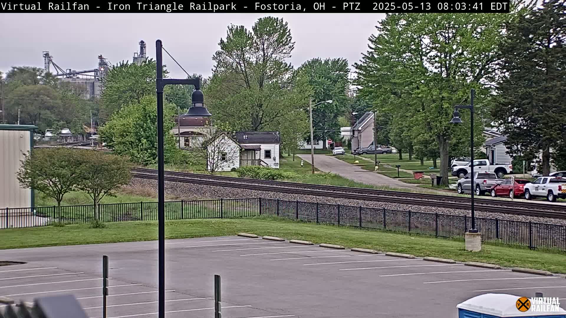 A view of railroad tracks beside a residential street on an overcast day, with a parking lot and industrial buildings in the background.