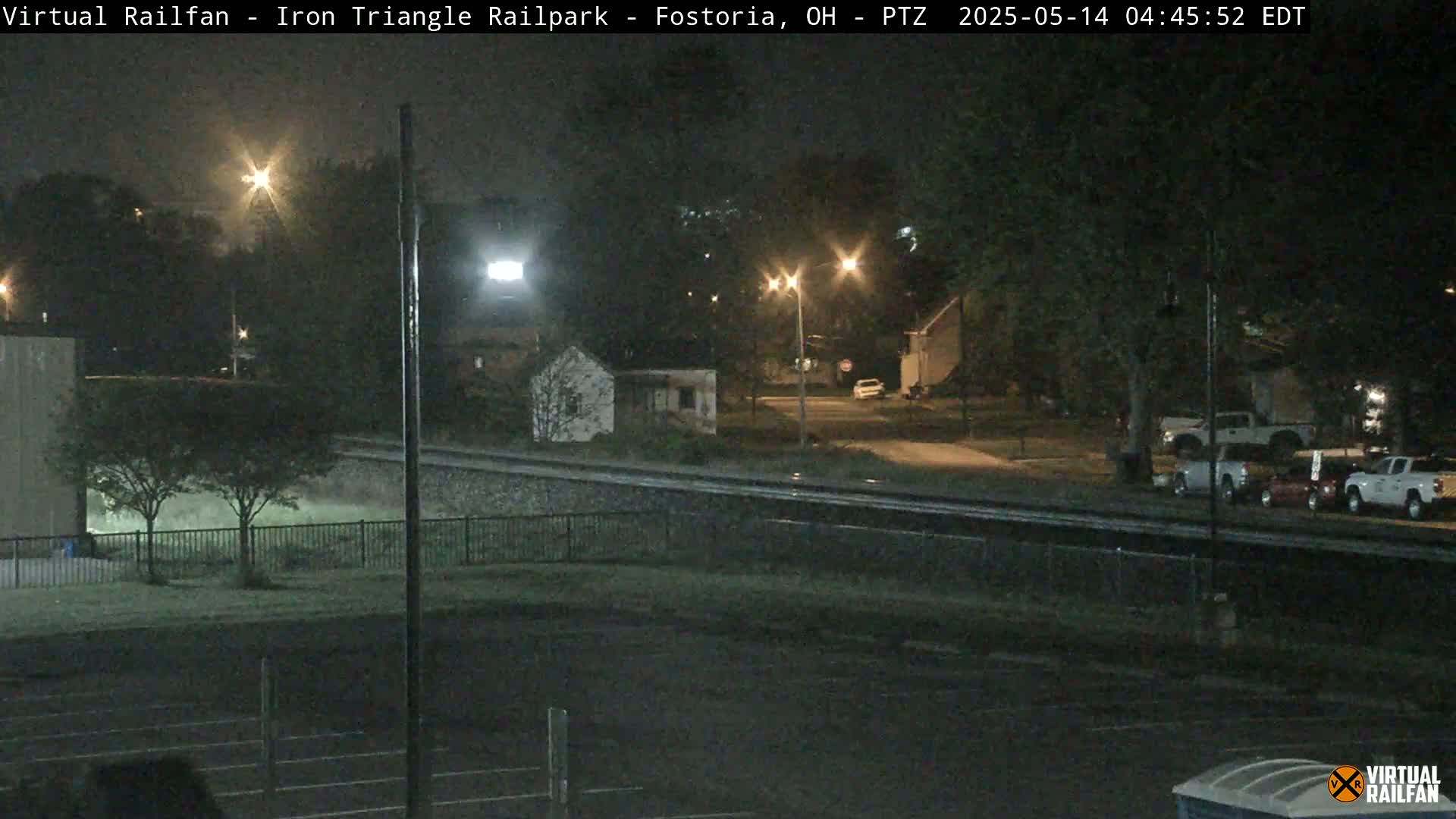 A nighttime view of a railroad crossing near a residential area, with several vehicles parked nearby under slightly overcast conditions.