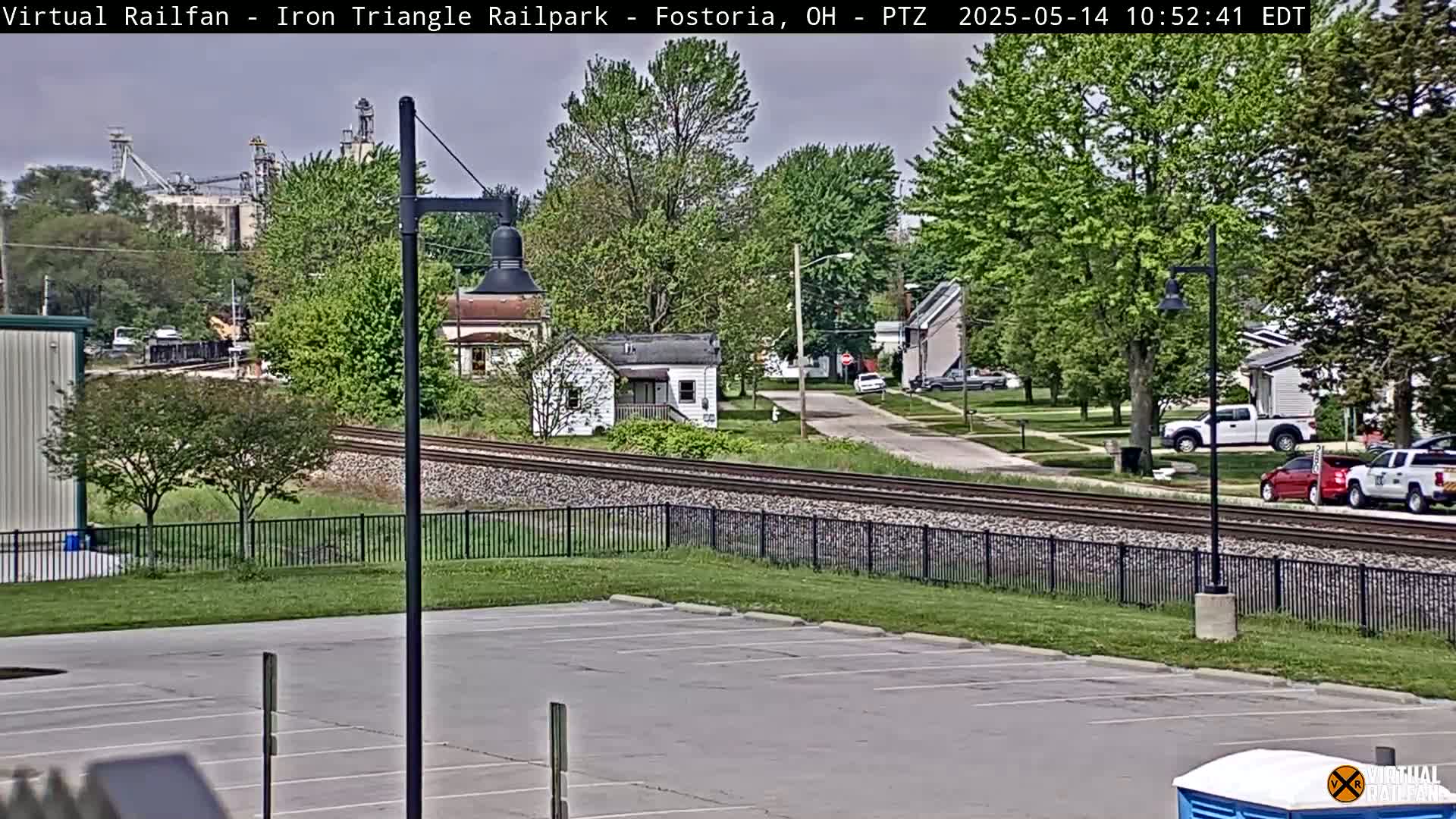 A mostly sunny view of railroad tracks bordering a residential area with parked cars, a few houses, and an empty parking lot.