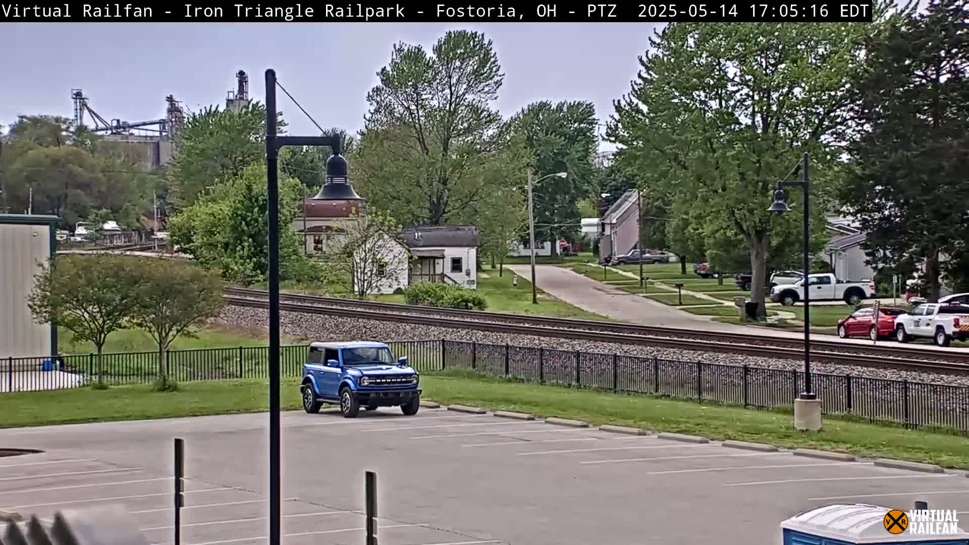 A blue SUV is parked in an empty lot next to railroad tracks, with a residential street and industrial buildings visible in the background on an overcast day.