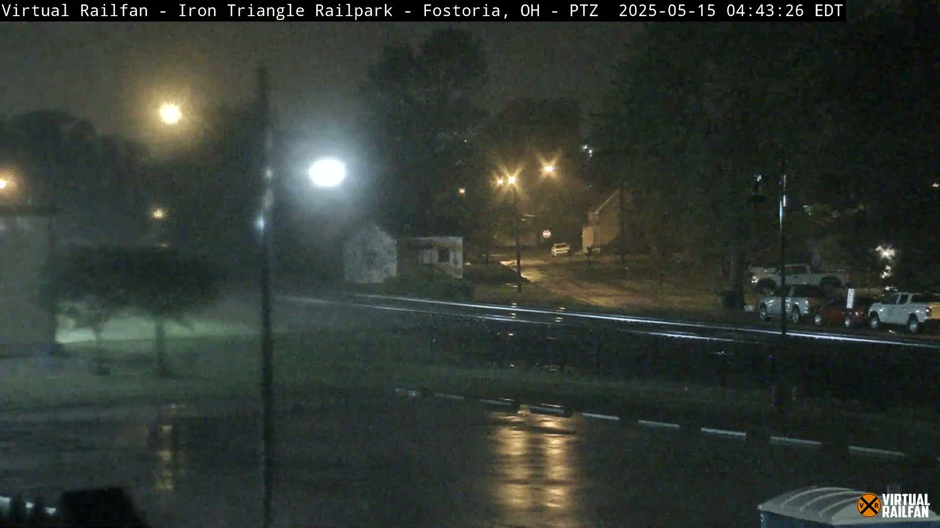 A nighttime, rainy scene shows a wet roadway, parked vehicles, and streetlights illuminating trees and buildings.