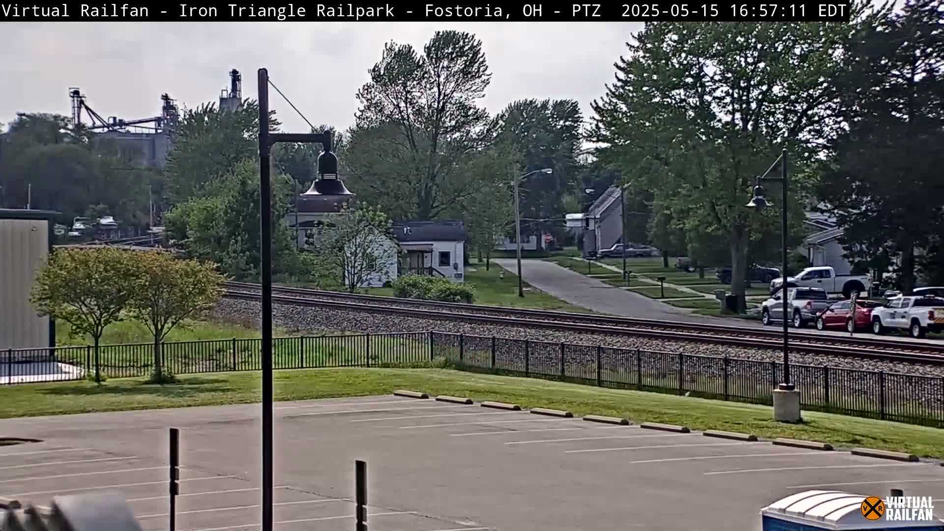 A mostly sunny day reveals a view of railroad tracks separating a parking lot from a residential street and industrial buildings in the distance.