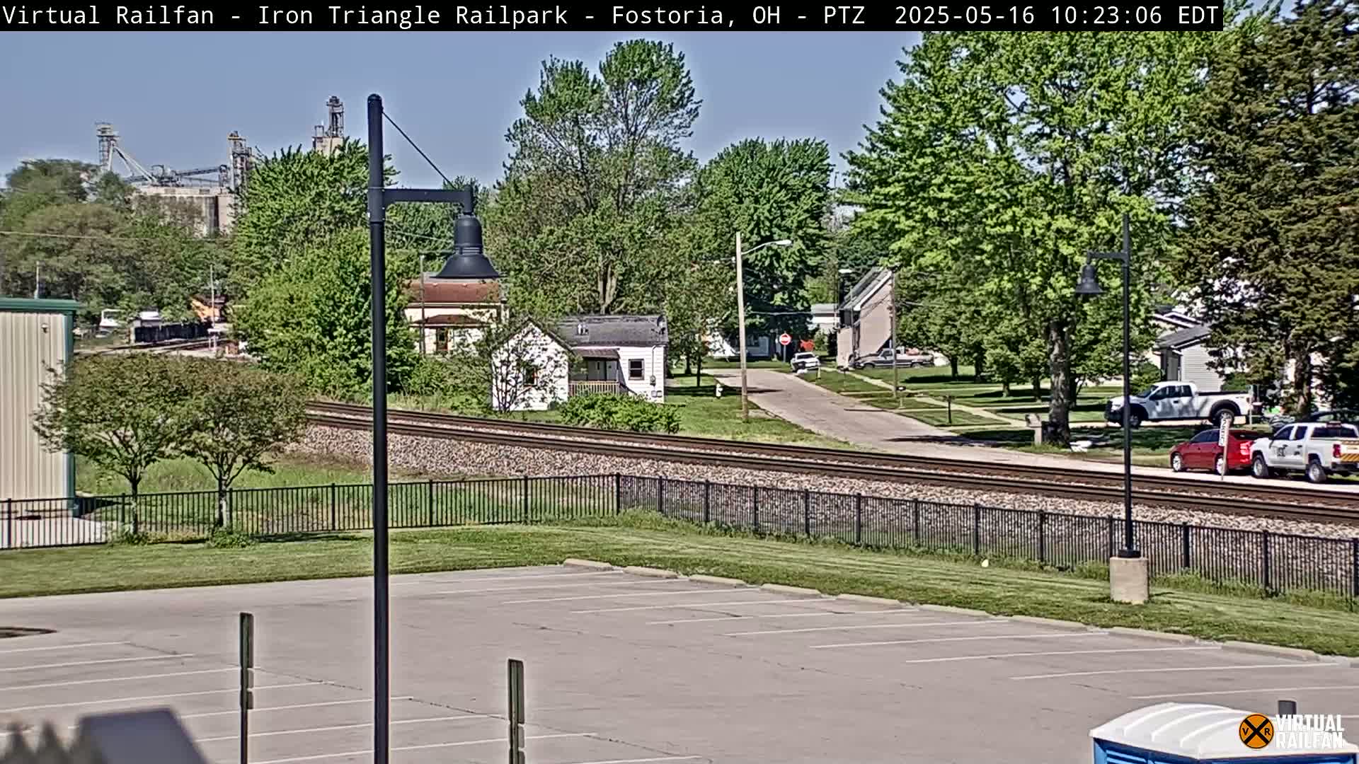 A mostly sunny view of a residential area with railroad tracks, a parking lot, and industrial buildings in the distance.