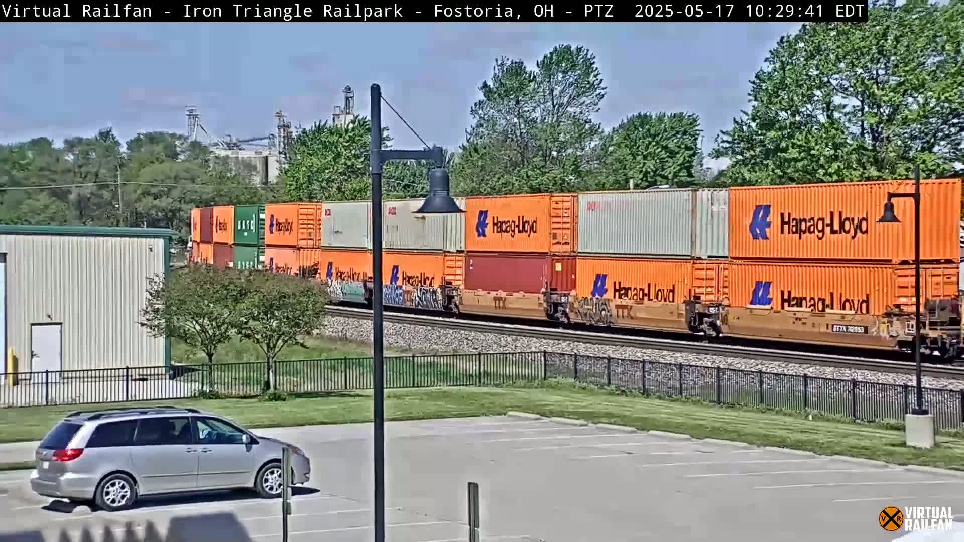 A silver minivan is parked in a lot next to a train carrying orange shipping containers under a partly sunny sky.