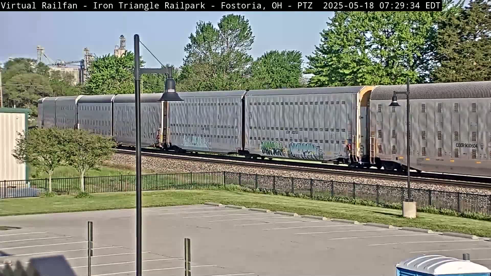 A long freight train with graffiti on some cars passes by a parking lot on a sunny day.