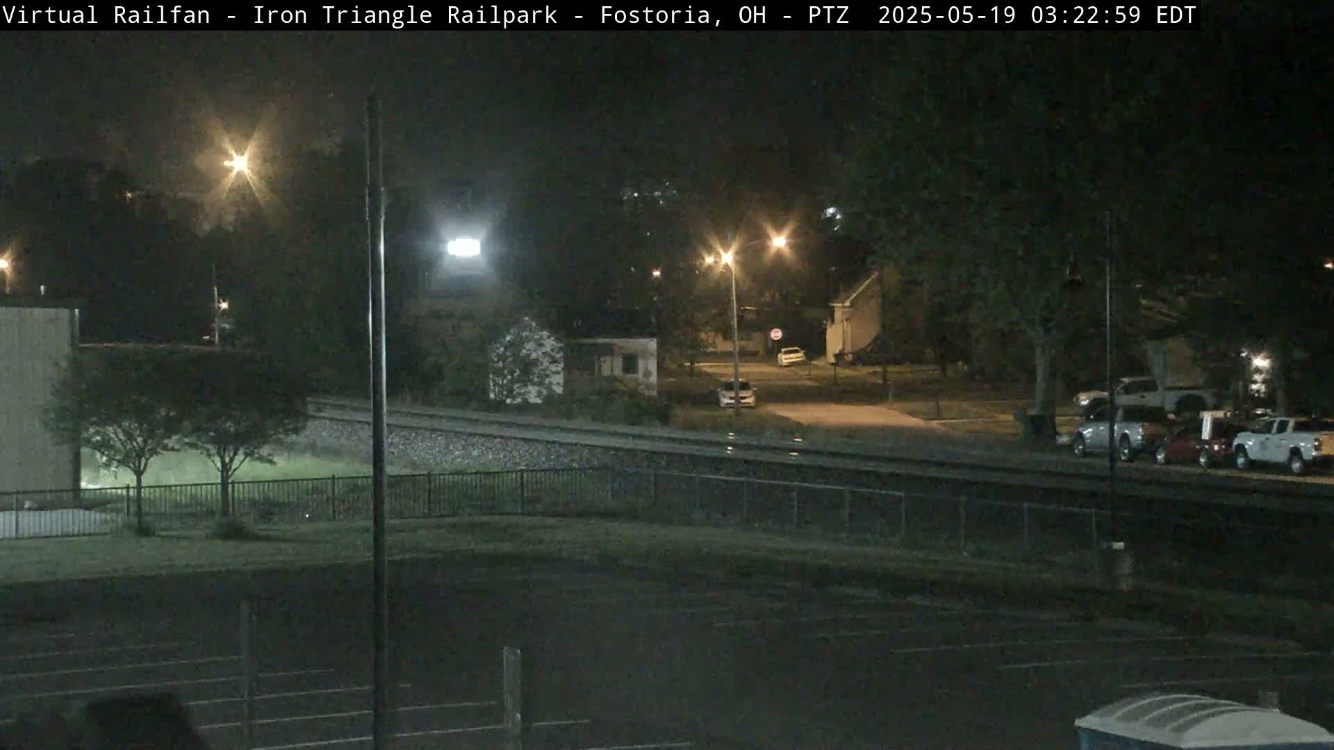 A nighttime view of a railroad track near a residential area, with several cars parked and streetlights illuminating the scene.