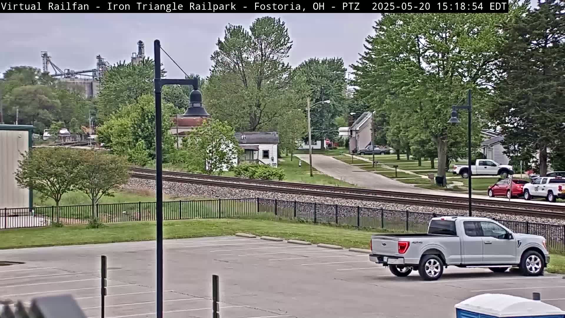 A mostly sunny day reveals a view of railroad tracks separating a parking lot with a silver pickup truck from a residential area with houses and trees, and a large industrial building in the background.