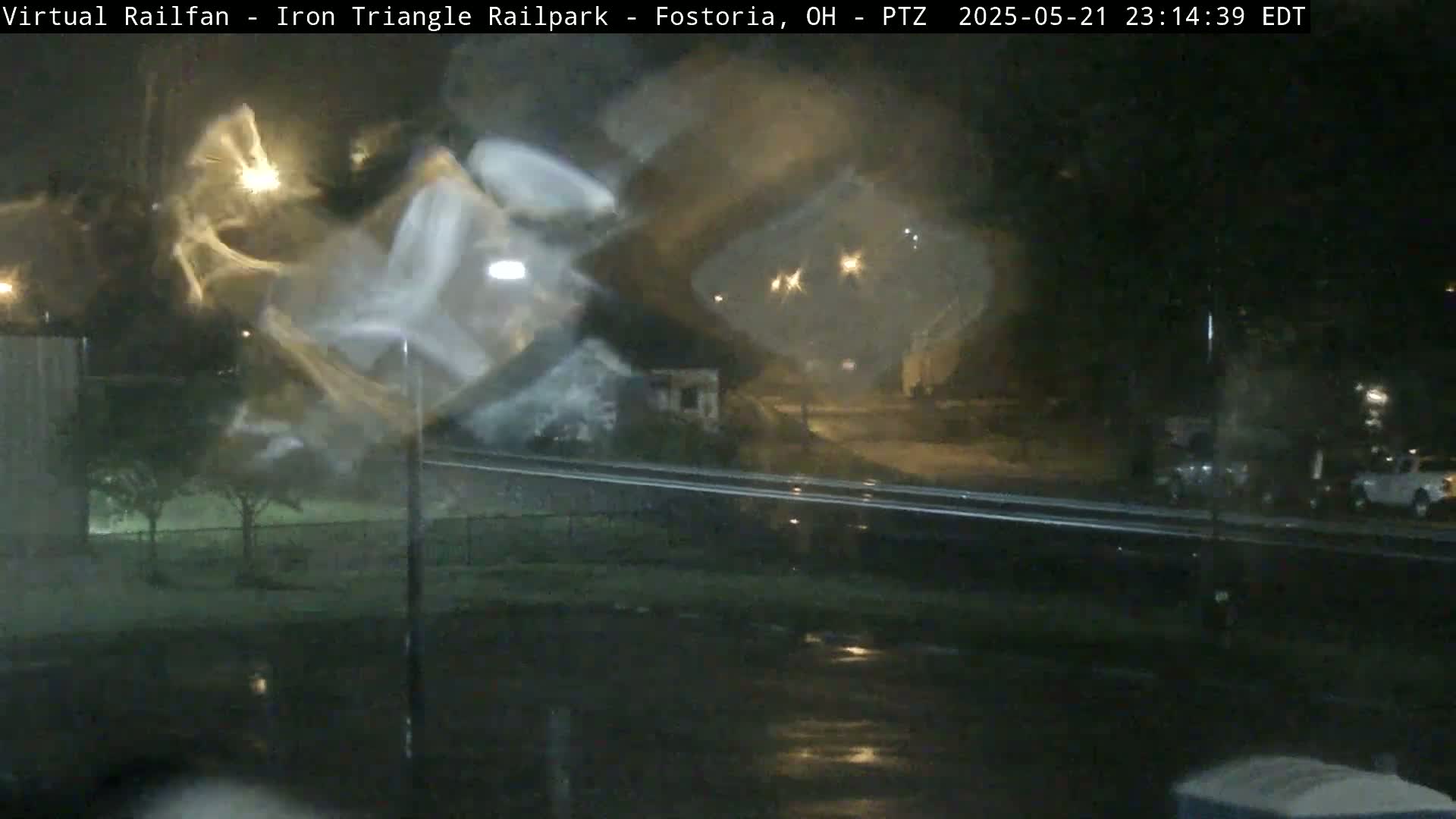 A rain-streaked window shows a nighttime view of a street scene with cars parked along the roadside and streetlights illuminating the wet pavement.