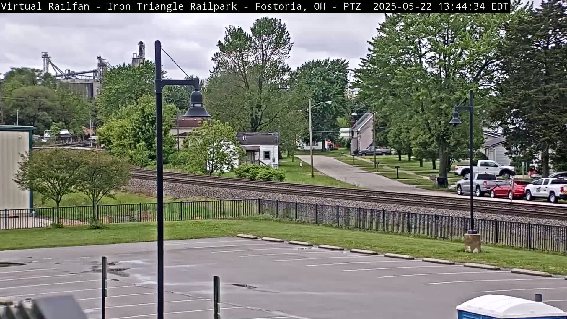 A mostly cloudy day shows a parking lot, a railroad track, several houses, and some industrial buildings in the distance.