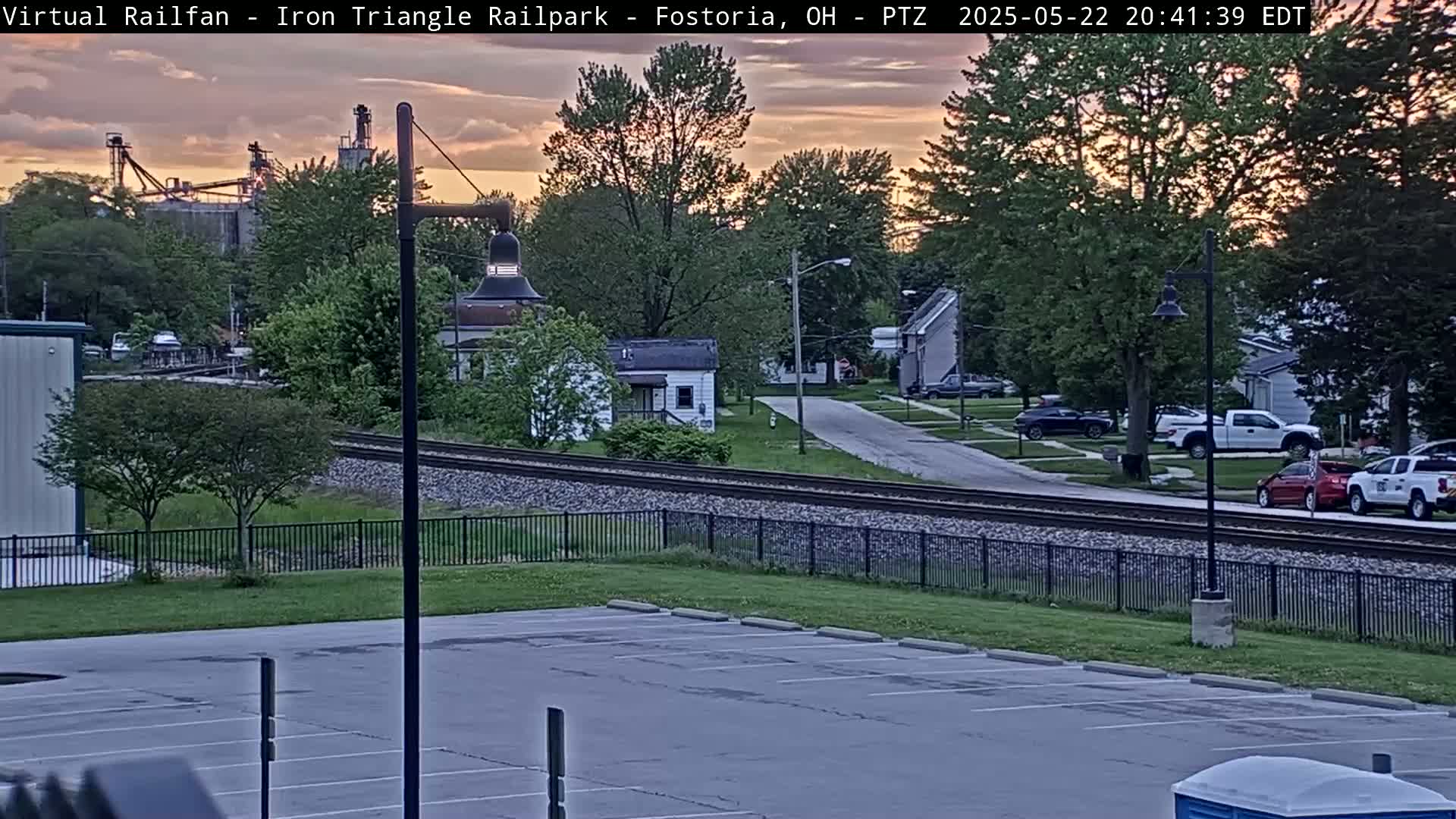 A parking lot overlooks a residential street and railroad tracks under a sunset sky.