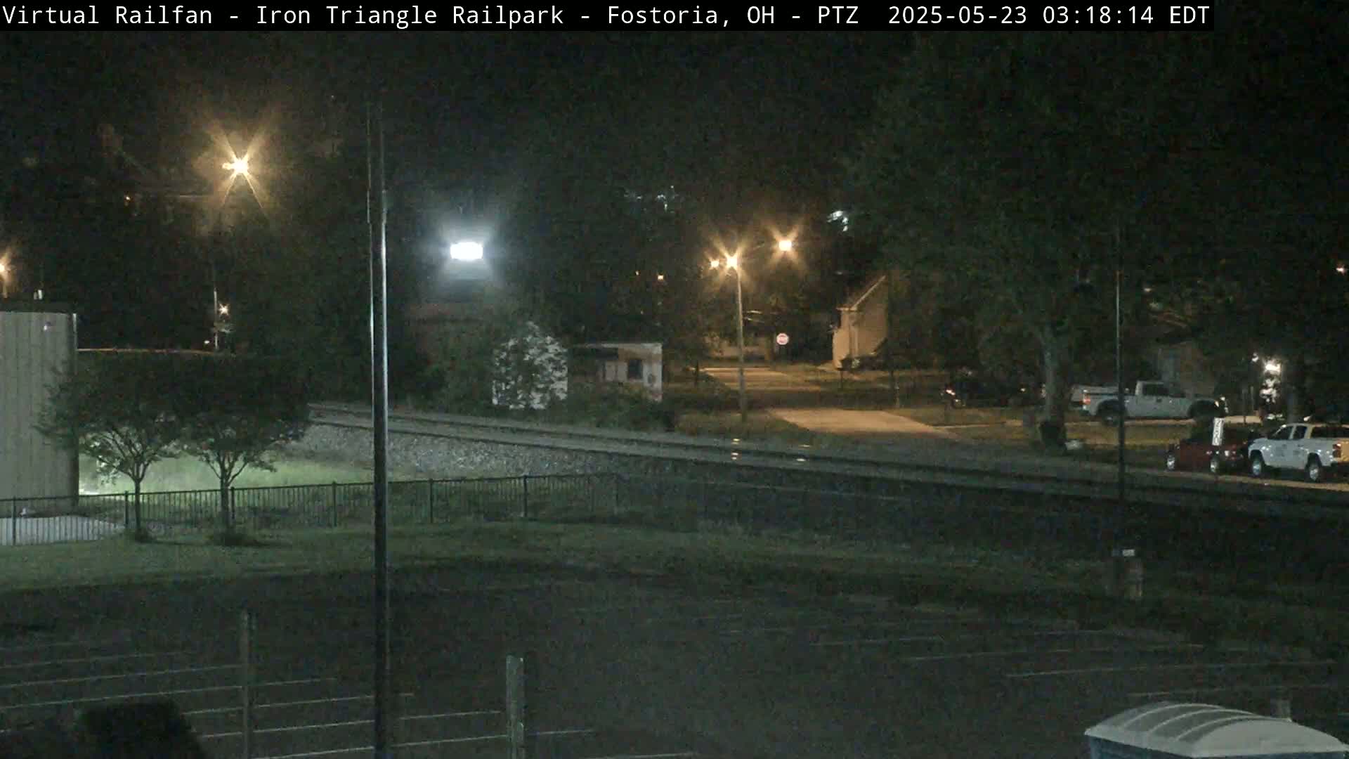 A nighttime view of a rail yard near a residential area, illuminated by streetlights, with several vehicles parked nearby.