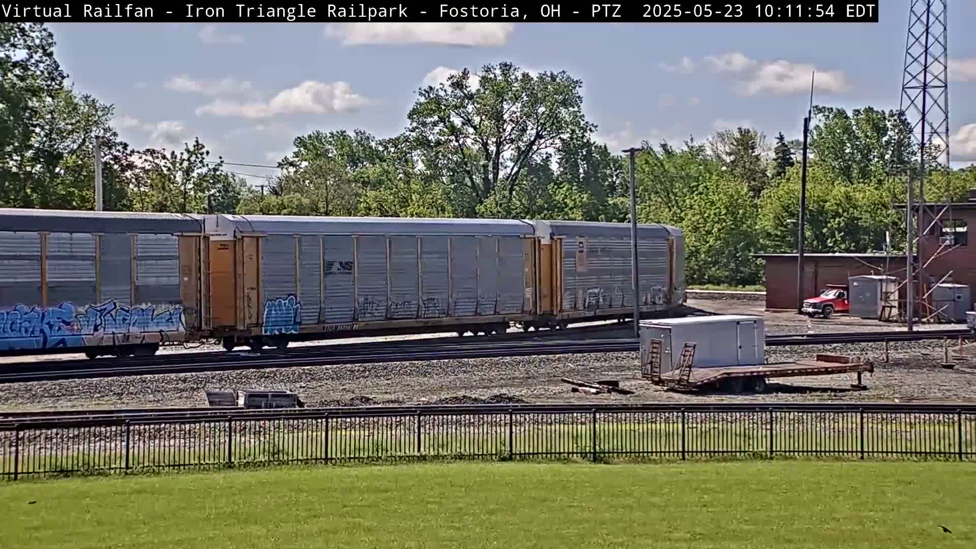 A partly cloudy day shows a graffiti-covered freight train passing by a flatbed trailer and a red pickup truck near a fenced-in area.