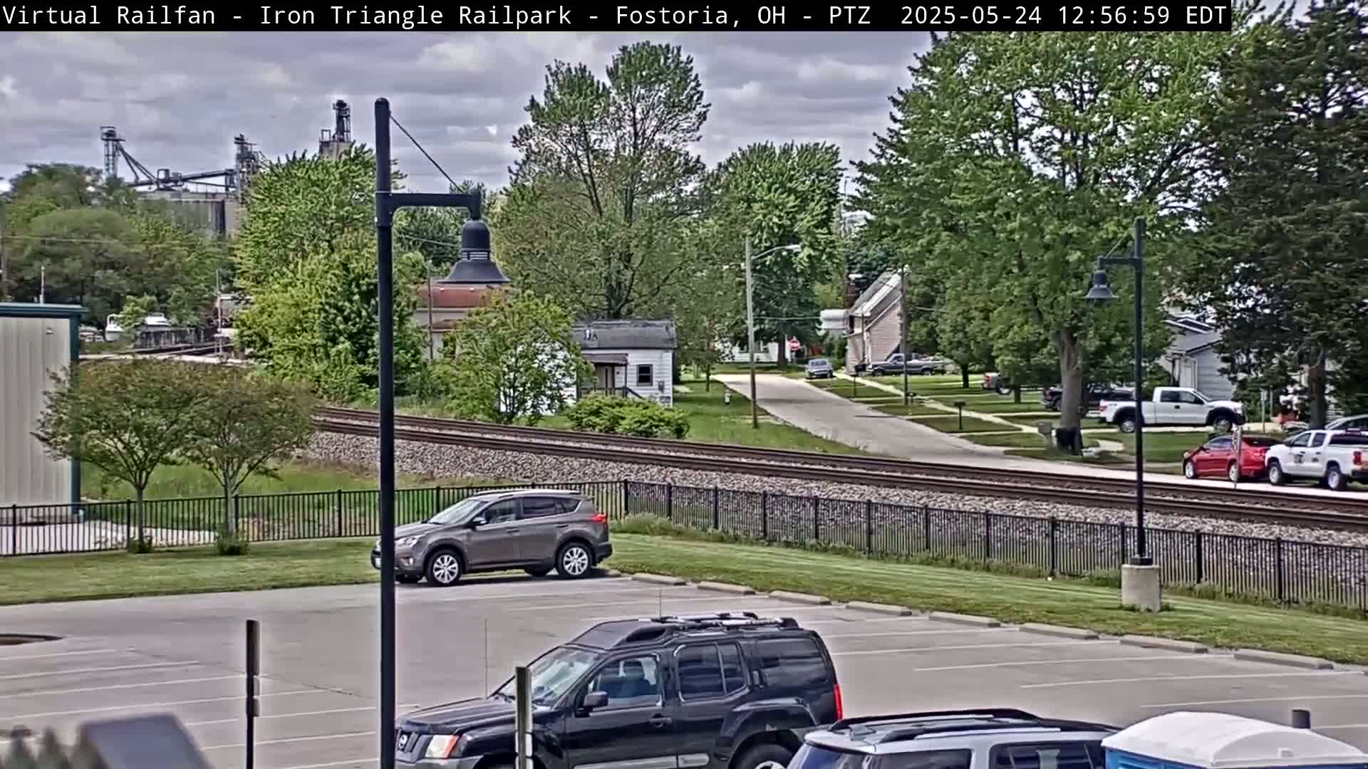 A partly cloudy day reveals a suburban scene with railroad tracks running through it, several cars in a parking lot and parked on the street, and industrial buildings in the distance.
